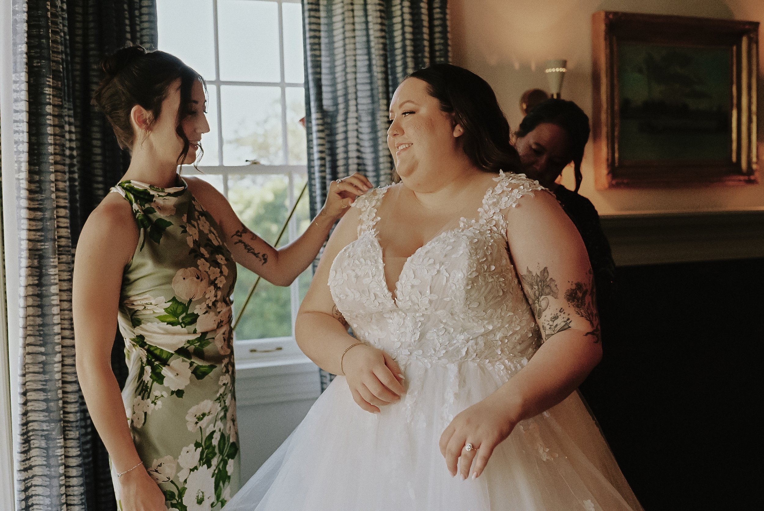 A bride in a white wedding dress smiling as she is helped by a woman in a floral dress, with another woman in the background, in a room with a window and a framed painting.