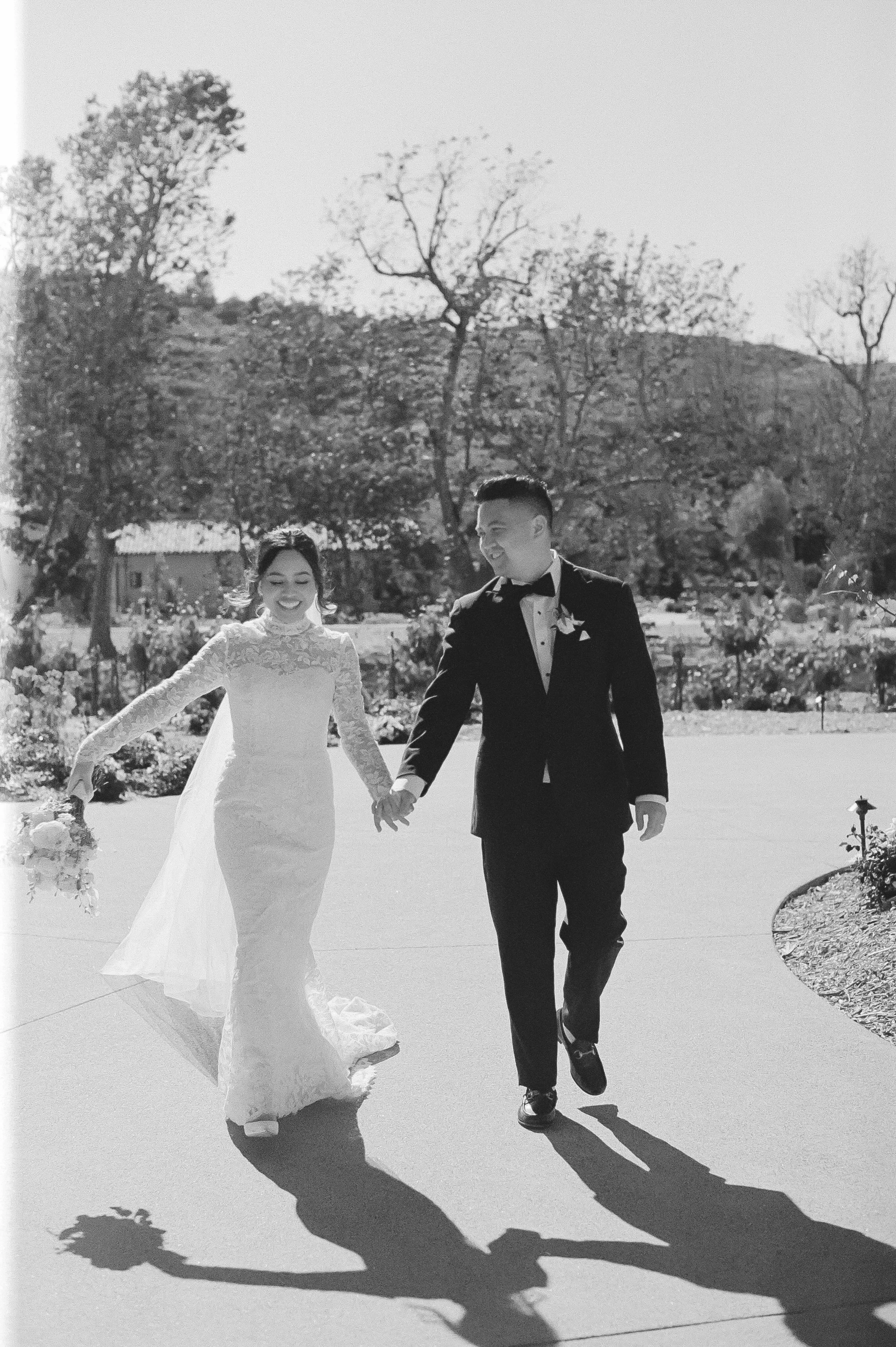 A bride and groom in wedding attire holding hands and walking outdoors on a sunny day, smiling.