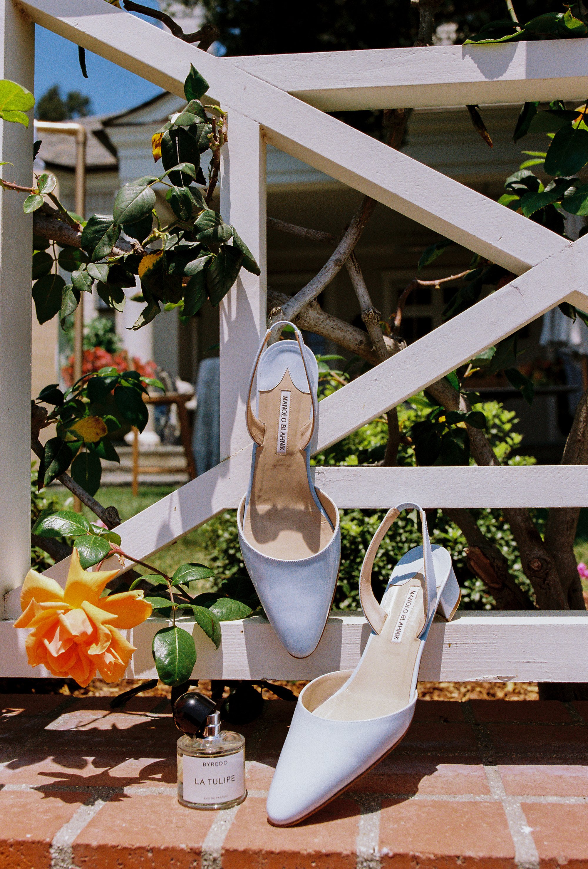 Pair of white high-heeled shoes hanging on a white wooden fence outdoors, with a small bottle of perfume labeled 'BYREDO LA TULIPE' placed on brick surface below the shoes, and orange flowers with green leaves nearby.