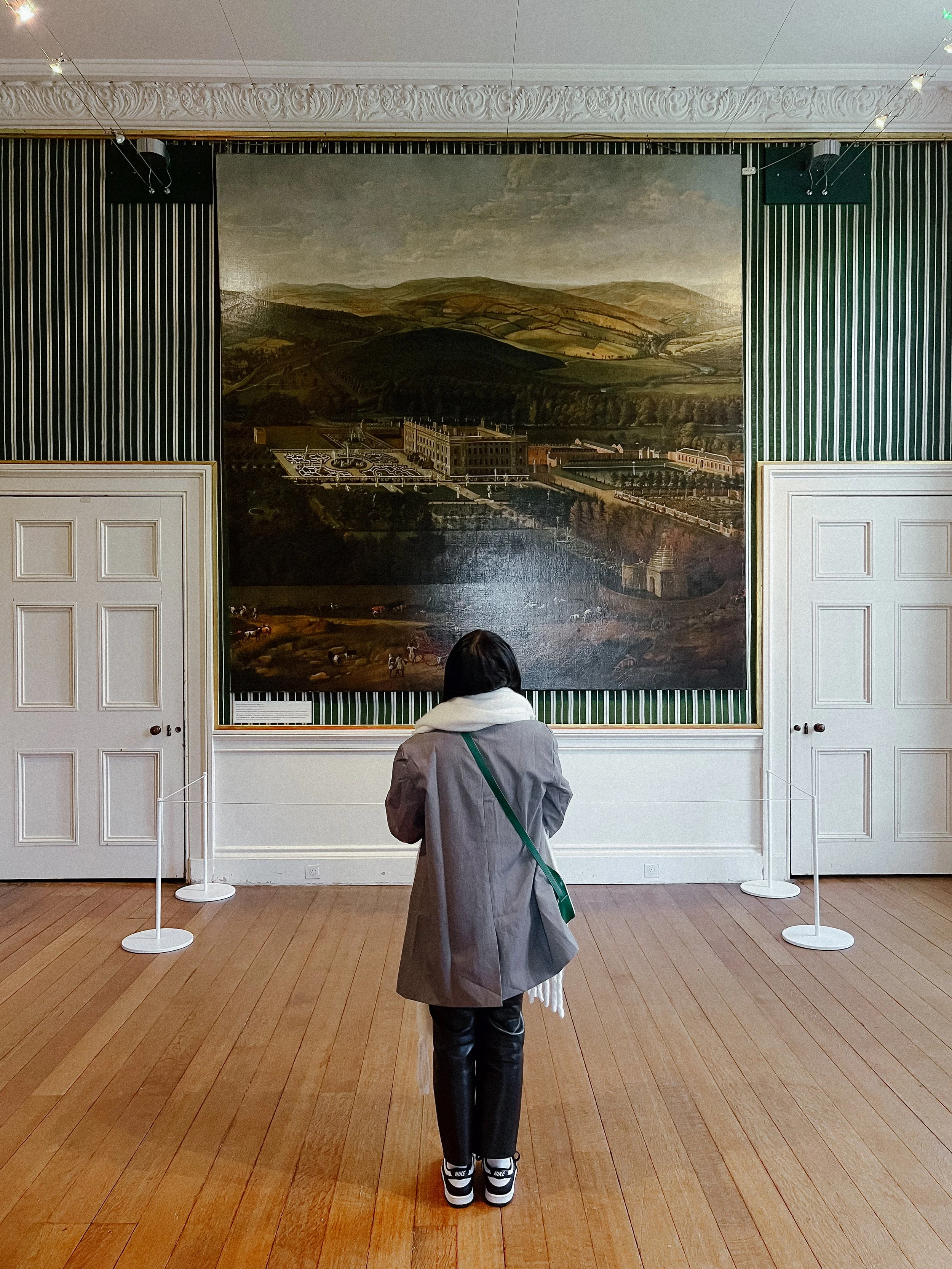 A person with a gray coat and black pants is standing in front of a large painting of a landscape in an art gallery. The painting depicts rolling hills, a building, and a cloudy sky.