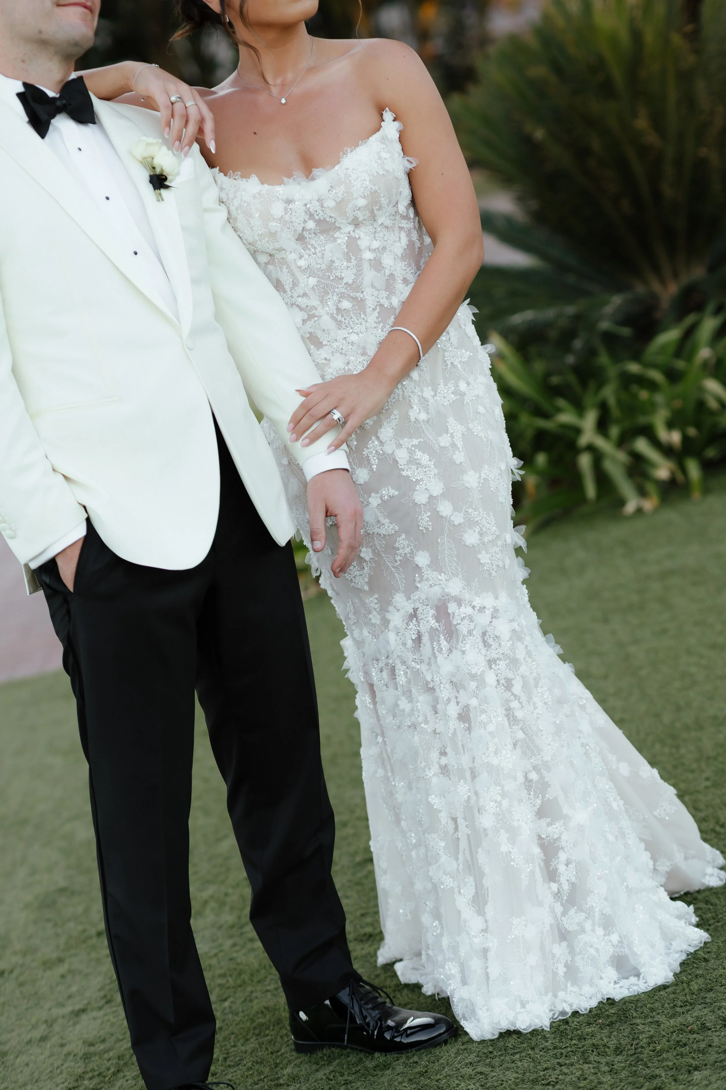 A bride and groom posing at their wedding, with the bride wearing a white lace wedding gown and the groom in a white tuxedo jacket with black pants and shoes.