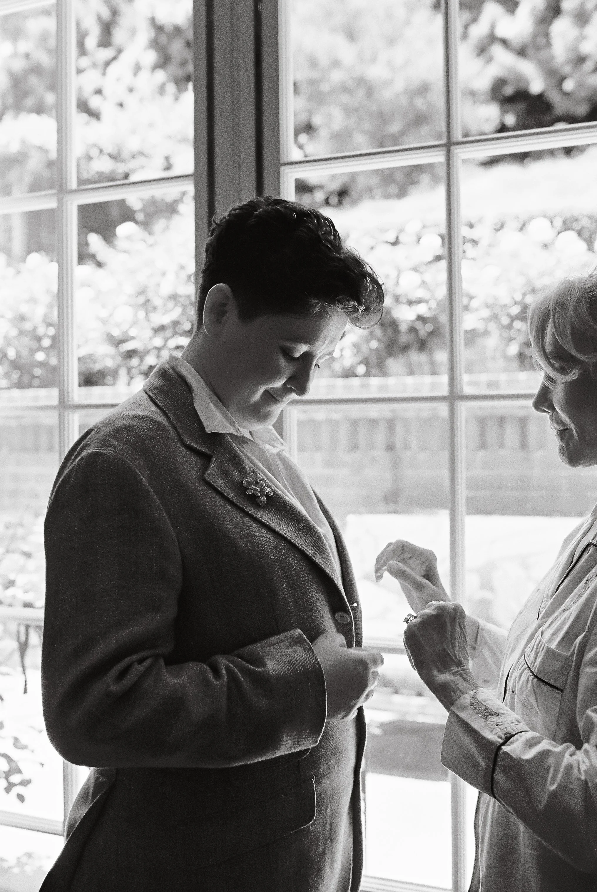 A black and white photo of two women, one younger and one older, standing near a large window. The younger woman is looking down and smiling, while the older woman is pinning something onto her jacket.
