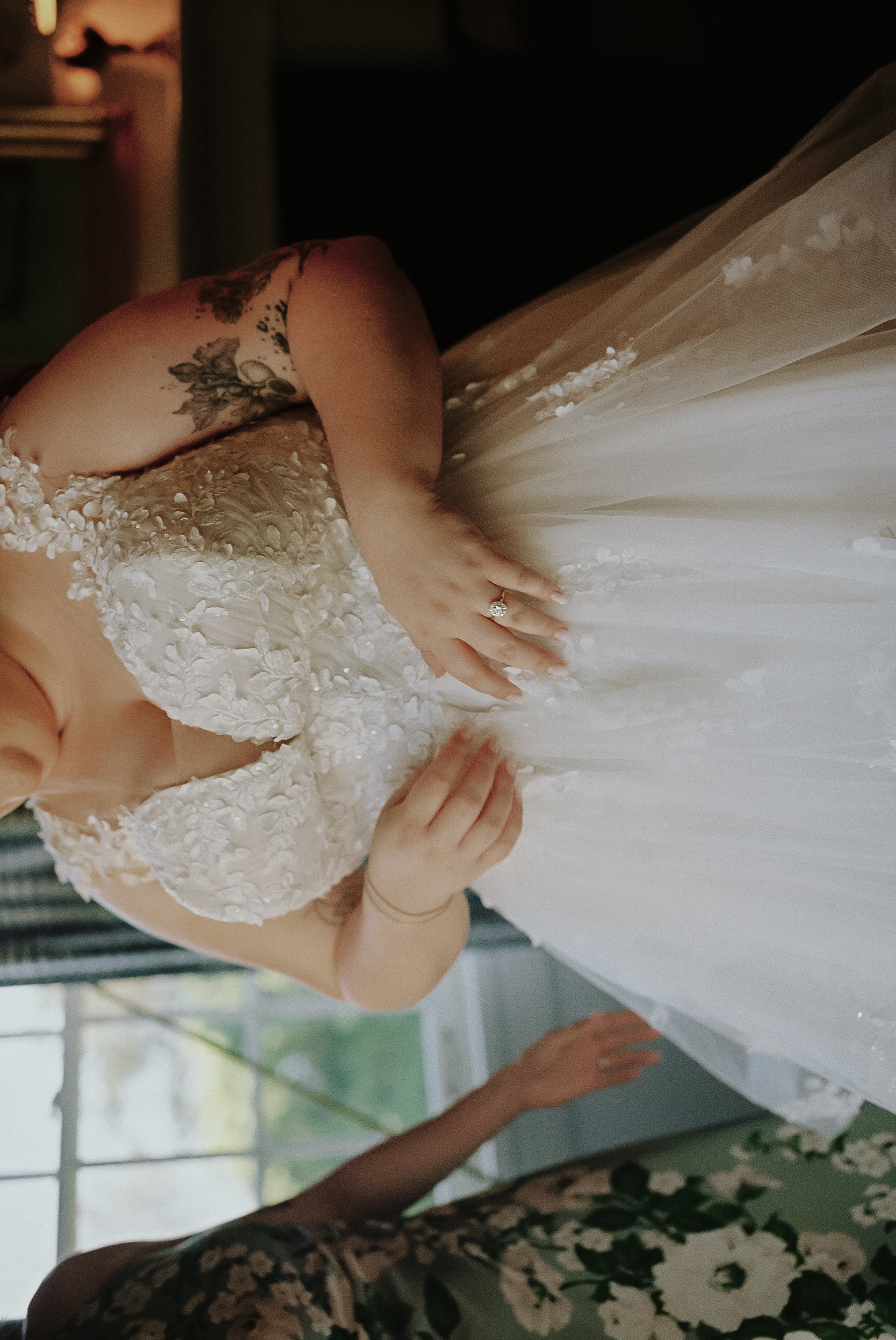Close-up of a bride in a wedding dress with floral embroidery, holding the fabric of her gown, showing her hand with an engagement ring, and a tattoo on her shoulder. Sunlight streams through a window in the background.
