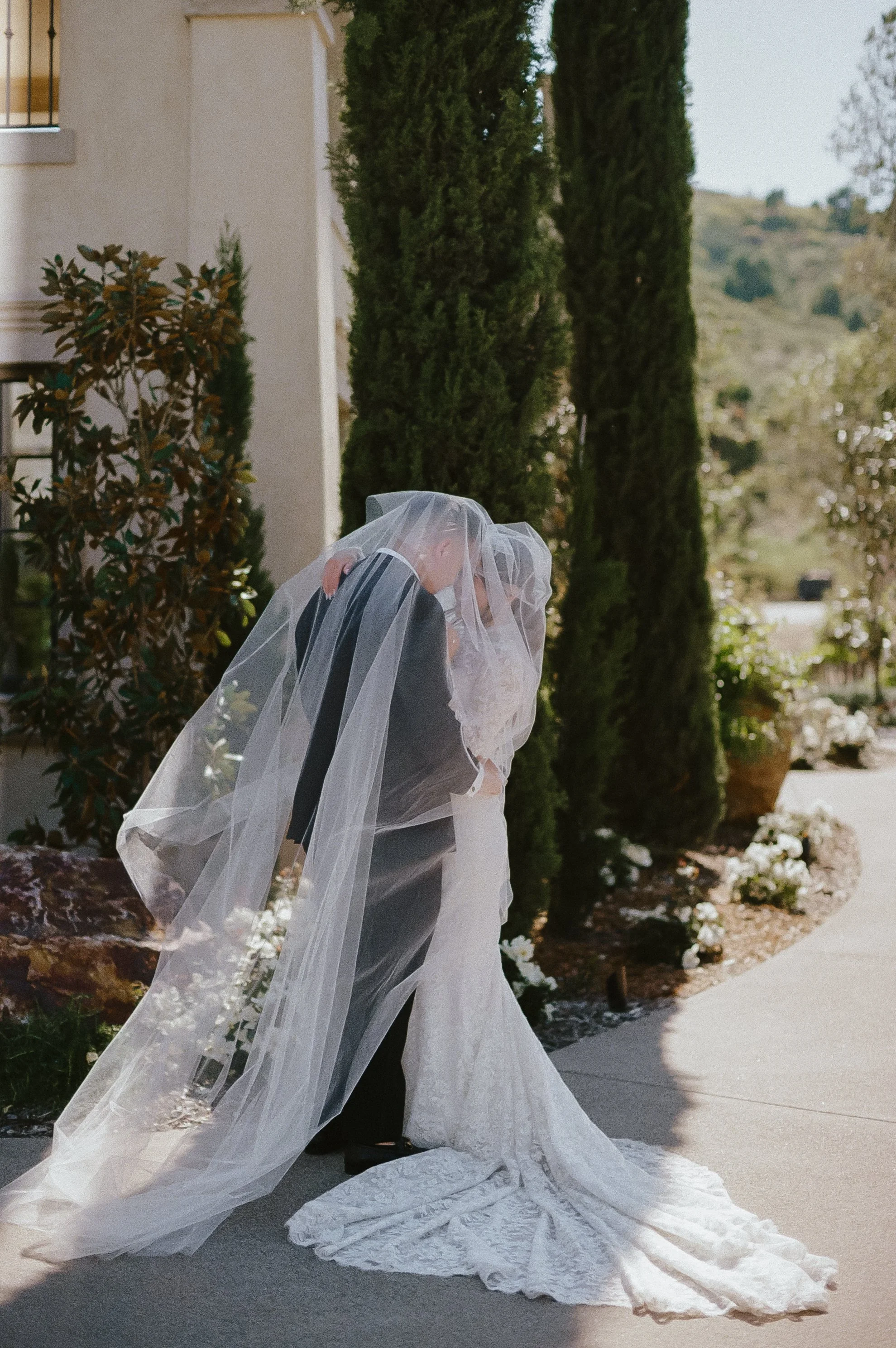 A bride and groom sharing a kiss outdoors, with the bride wearing a white lace wedding gown and veil, and the groom in a black suit, beneath tall green trees and in front of a beige building.