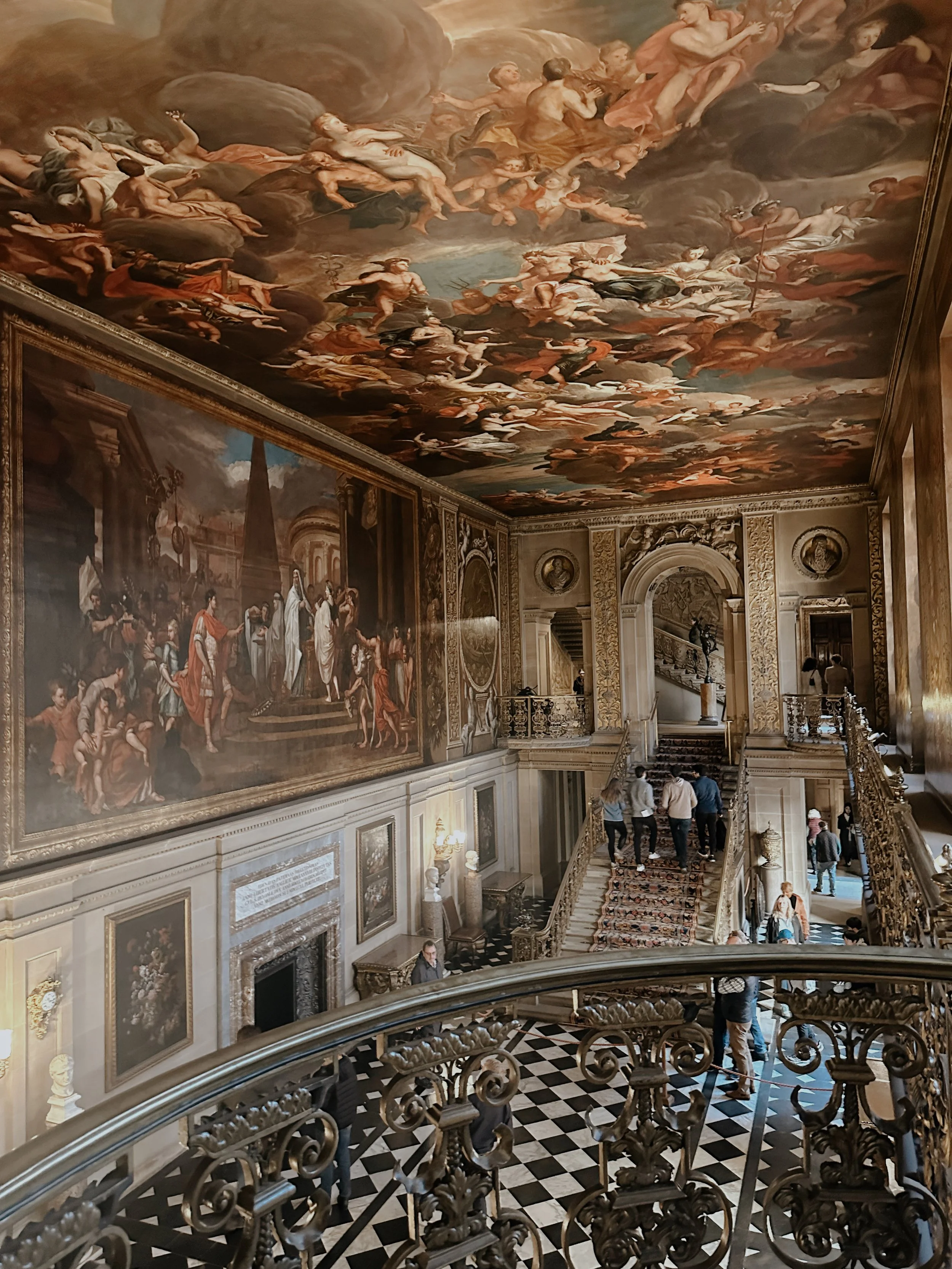 Interior of a grand, ornate room with a large painted ceiling, multiple large paintings, and a decorative staircase with visitors walking up and down.