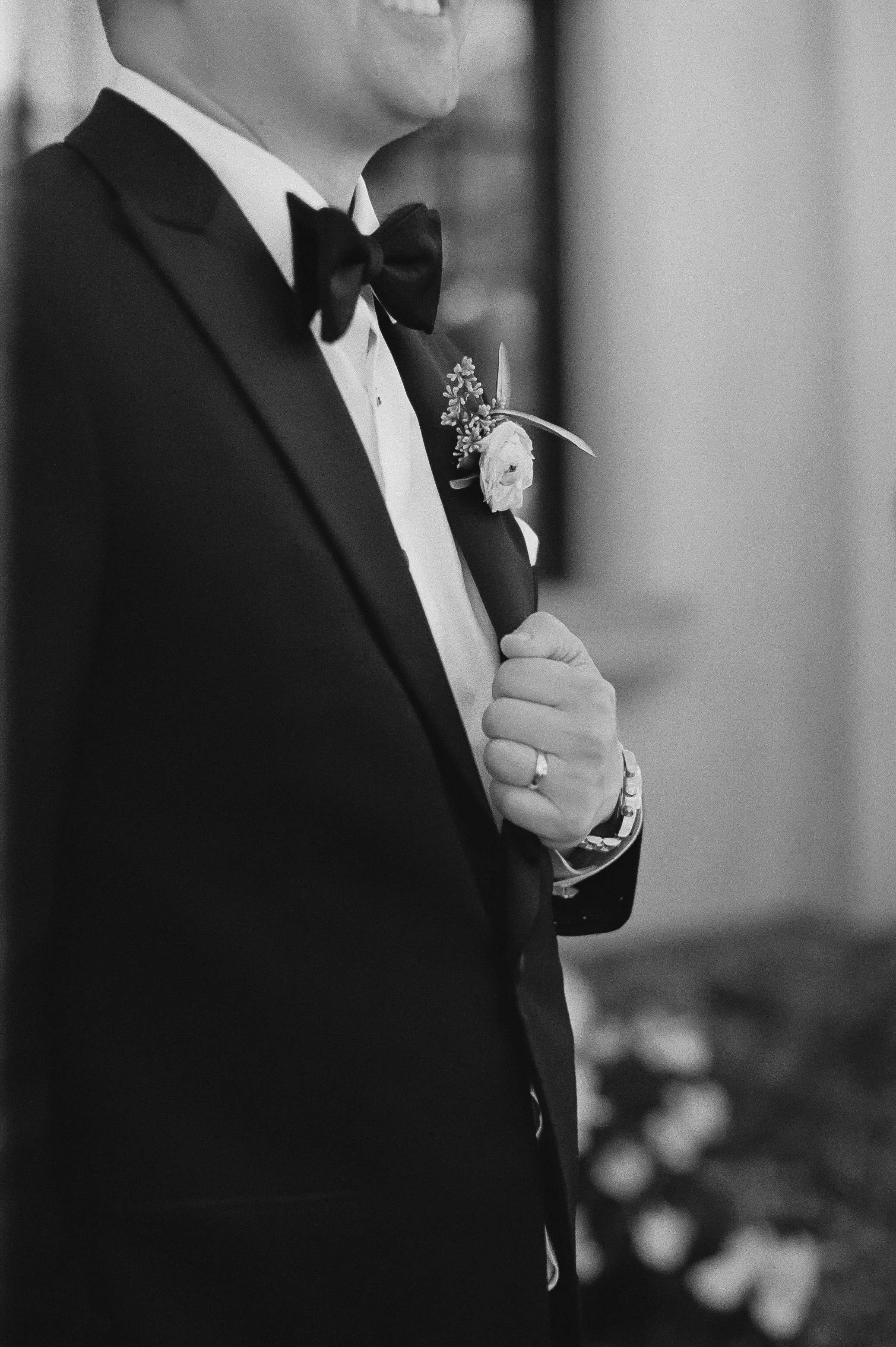 A man dressed in a tuxedo with a bow tie, holding his lapel with one hand that has a wedding ring and a wristwatch. The image is in black and white, with a blurred background, possibly at a wedding or formal event.