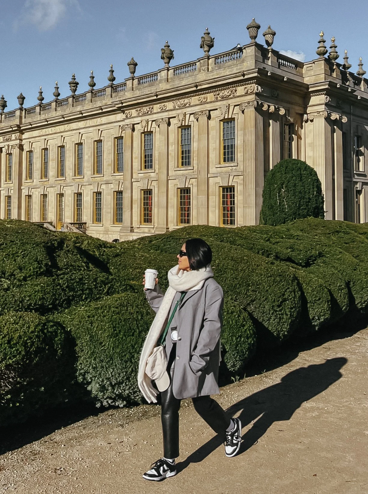 A woman with black hair, sunglasses, a gray coat, black pants, and sneakers walking outdoors with a coffee cup in hand, in front of a historic building with columns and decorative elements.