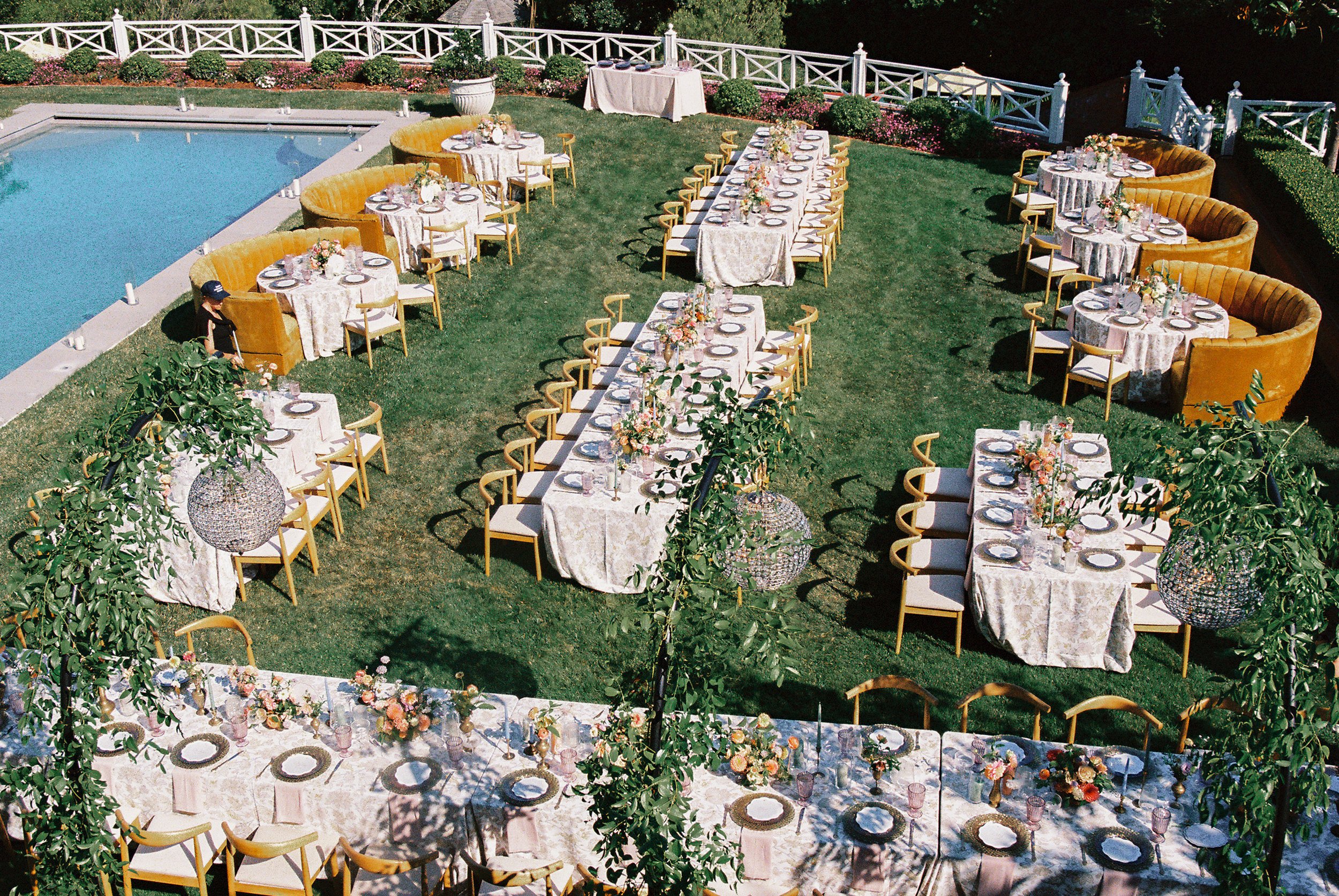 Outdoor garden wedding reception setup with long and round tables arranged on a grassy area, decorated with floral centerpieces, set with plates and glassware, surrounded by yellow chairs and large yellow velvet lounge seating near a swimming pool, w