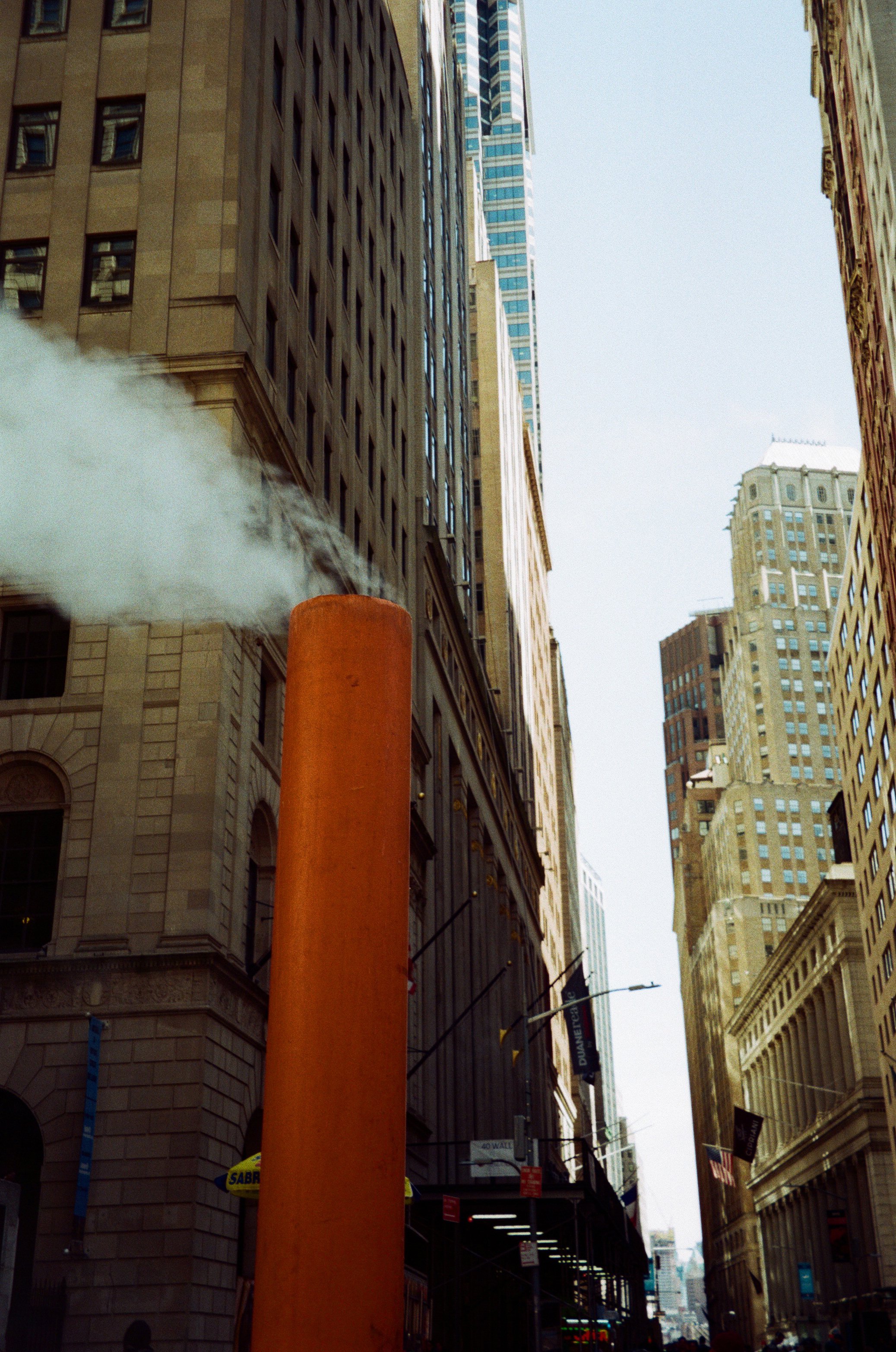 City street with tall buildings and a large orange chimney emitting white smoke or steam.