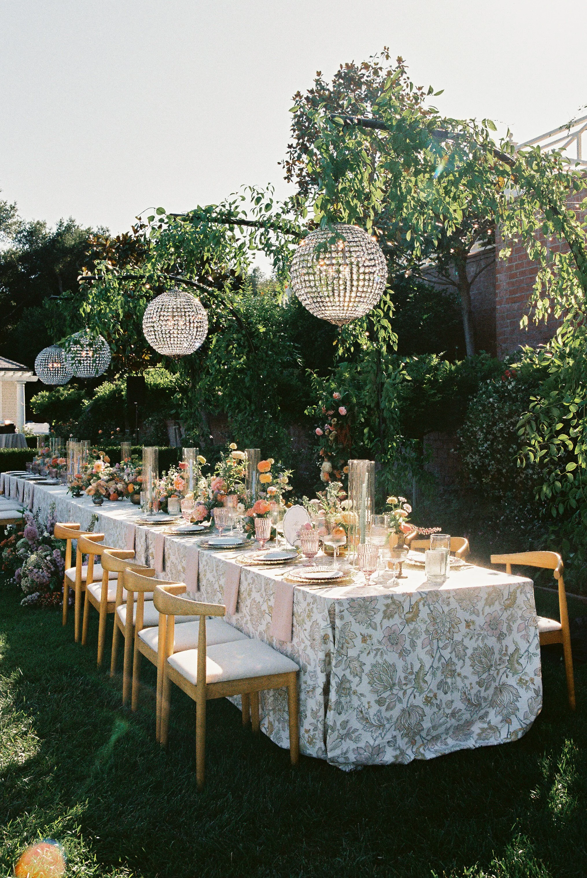 Outdoor wedding or specially decorated event setup with a long floral tablecloth, floral arrangements, glassware, and hanging spherical chandeliers, set on a grassy area with greenery and a brick building in the background.
