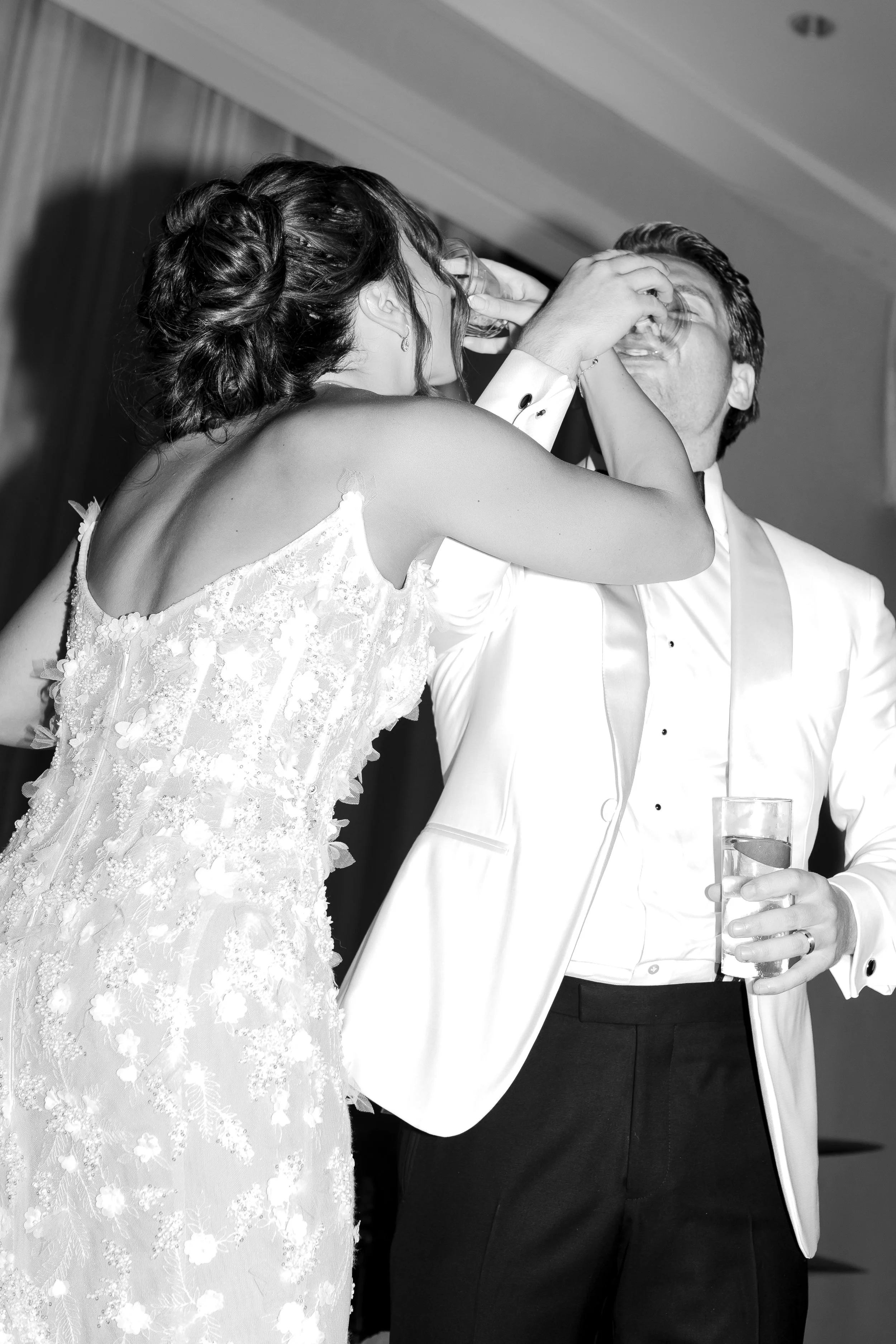 Black and white photo of a woman in a wedding dress feeding a man in a tuxedo cake or dessert at a celebration.