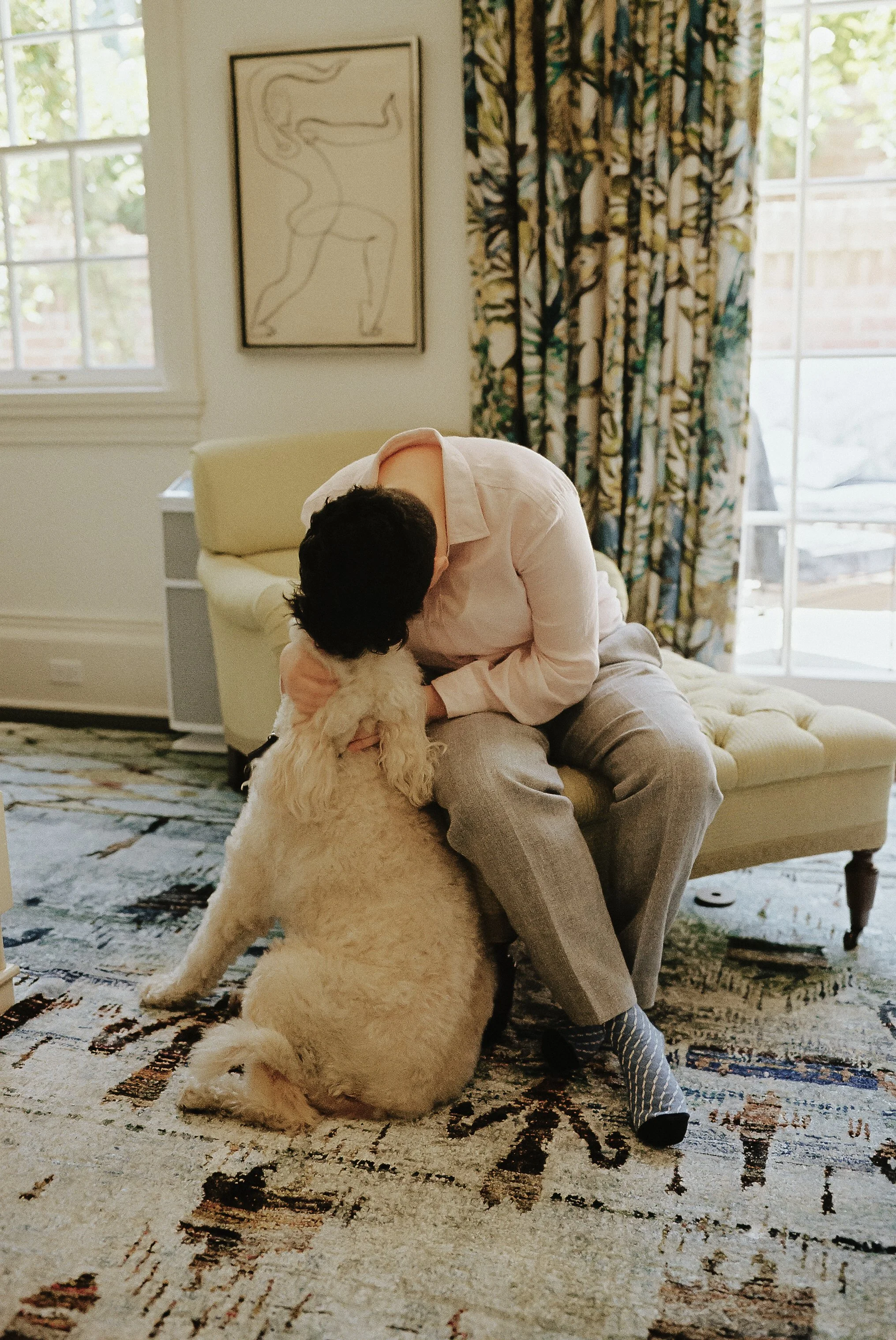 A man sitting on a yellow armchair, leaning forward and hugging a fluffy white dog, in a cozy living room with patterned curtains and artwork on the wall.
