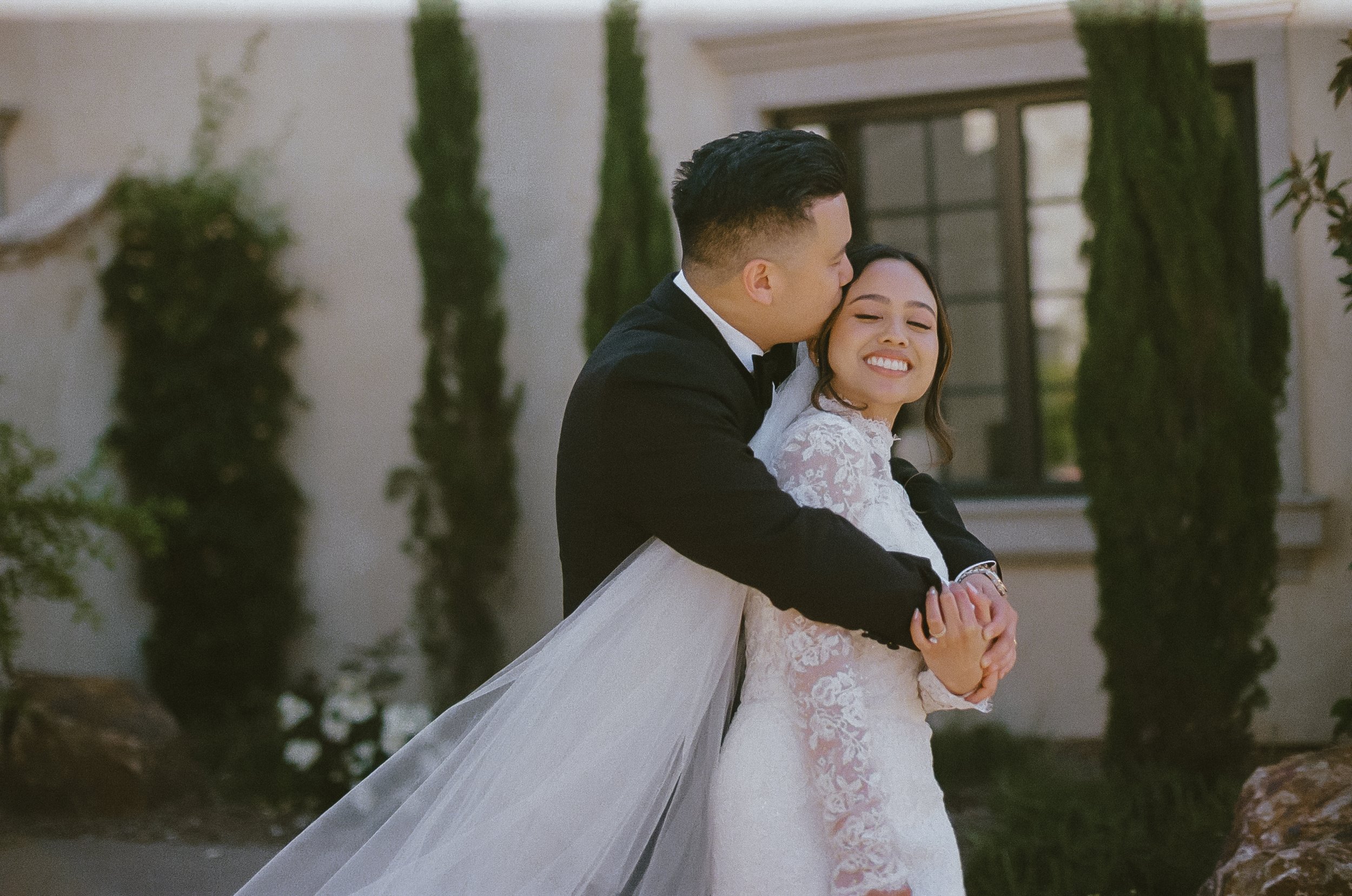 A happy bride and groom embracing outdoors during their wedding, with the groom kissing the bride on her temple and both smiling.