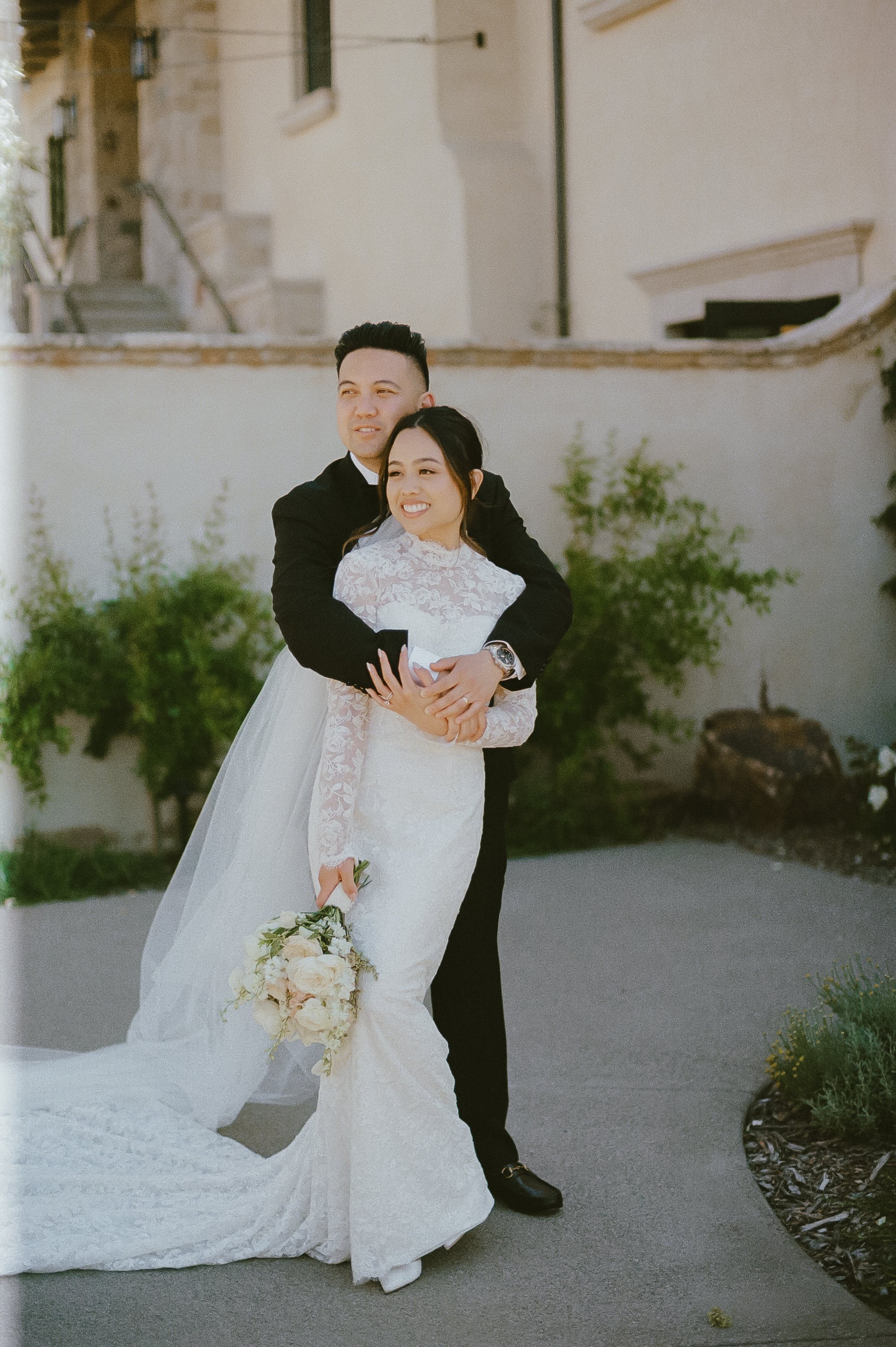 A bride and groom sharing a hug outdoors, the bride holding a bouquet of flowers, both smiling.