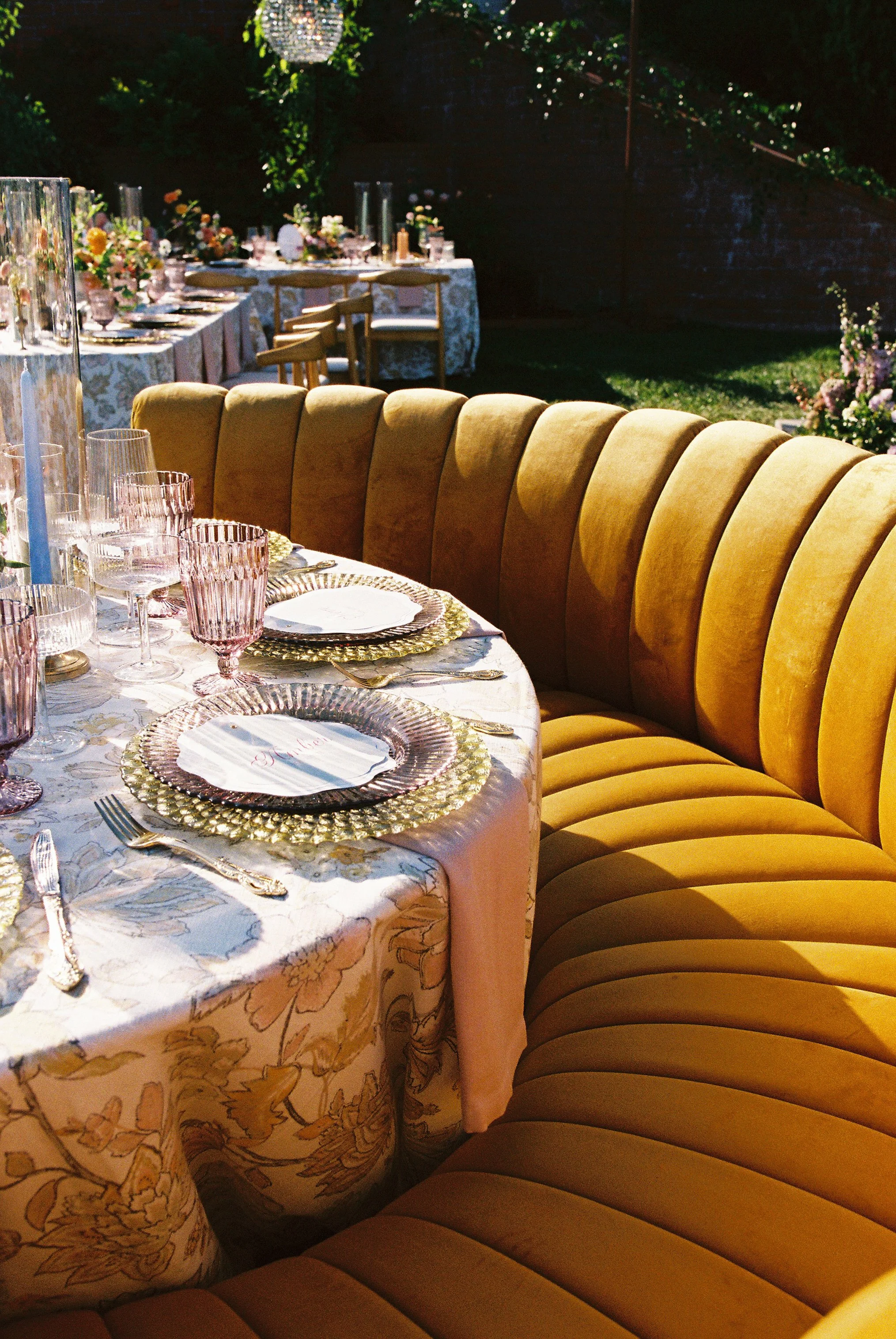 An elegant outdoor party setup with a long round dining table covered with a floral tablecloth, set with gold-rimmed plates, pink-tinted glassware, and gold utensils, surrounded by a curved, mustard-colored velvet sofa, with additional tables and flo