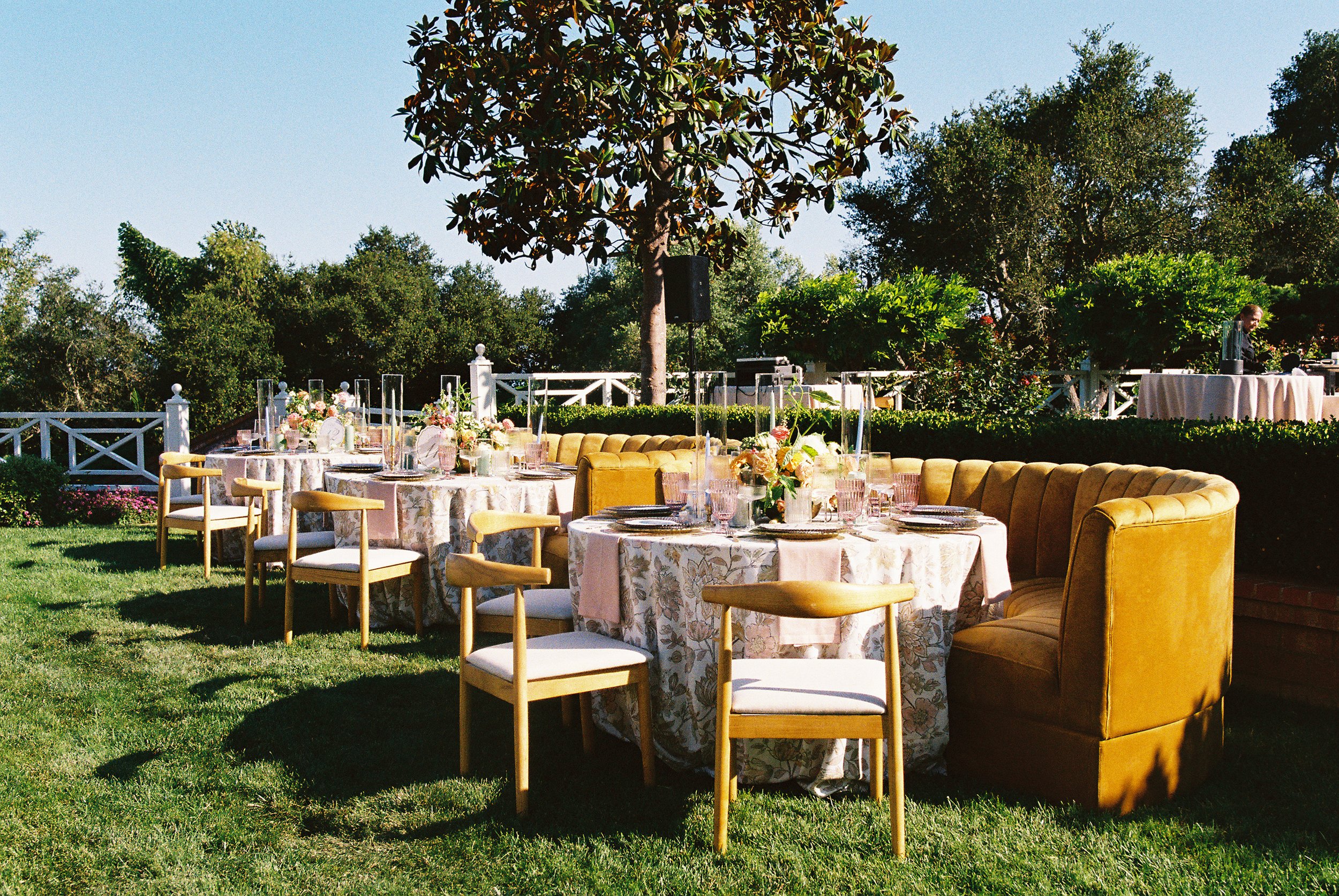 Elegant outdoor event setup with round tables decorated with floral tablecloths and centerpieces, surrounded by a mix of wooden chairs and a yellow velvet banquette, on a grassy lawn with trees and a white fence in the background.