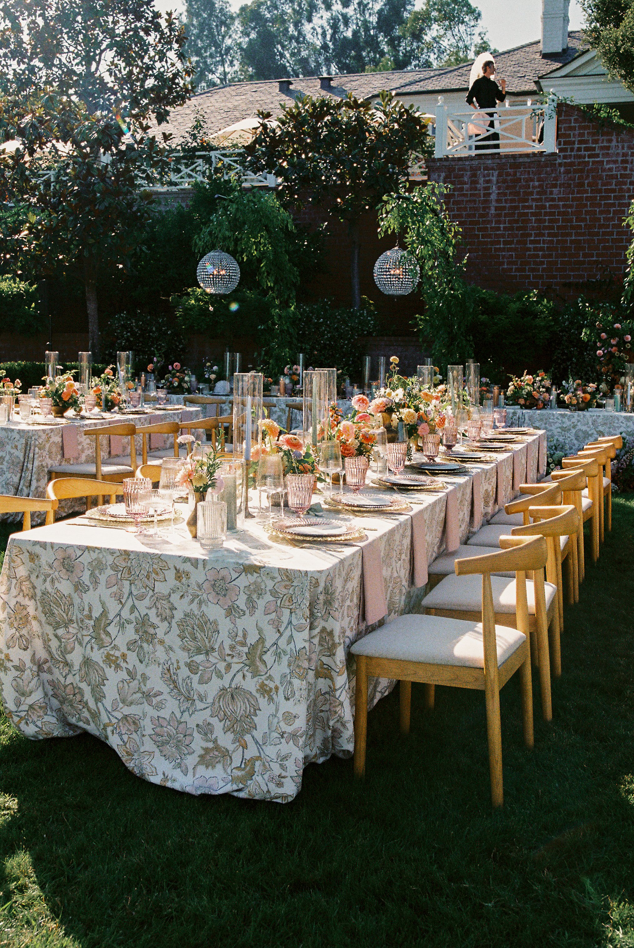 Outdoor wedding reception with tables covered in floral tablecloths, decorated with pink and white flowers, glassware, and candles. String lights and hanging disco balls are in the background, with a woman standing on a balcony above.
