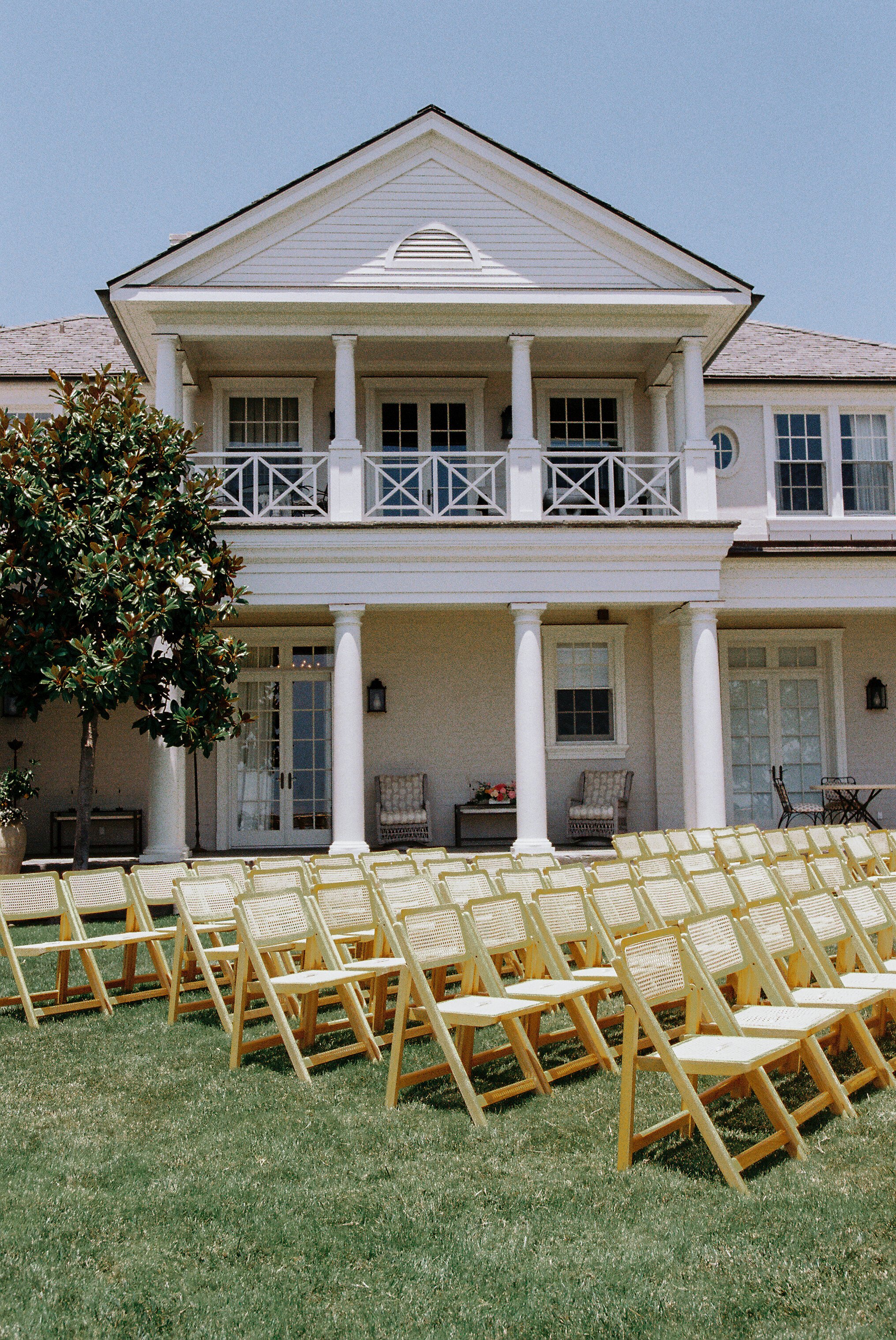 Outdoor wedding ceremony setup with rows of cream-colored folding chairs on a grassy lawn in front of a large, white, two-story house with a front porch and balcony, decorated with black lantern-style wall sconces, a table with chairs, and sitting ar