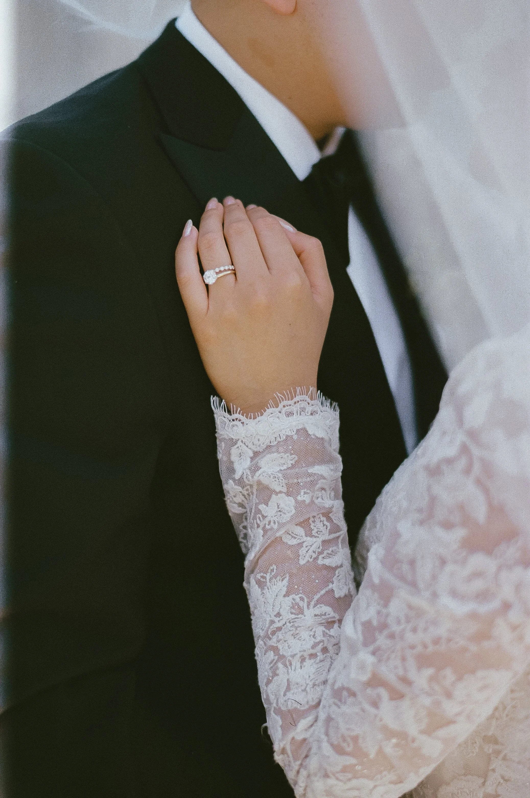 Close-up of a bride and groom with the bride's hand resting on the groom's chest, showing a wedding ring, lace wedding dress sleeve, and black tuxedo.