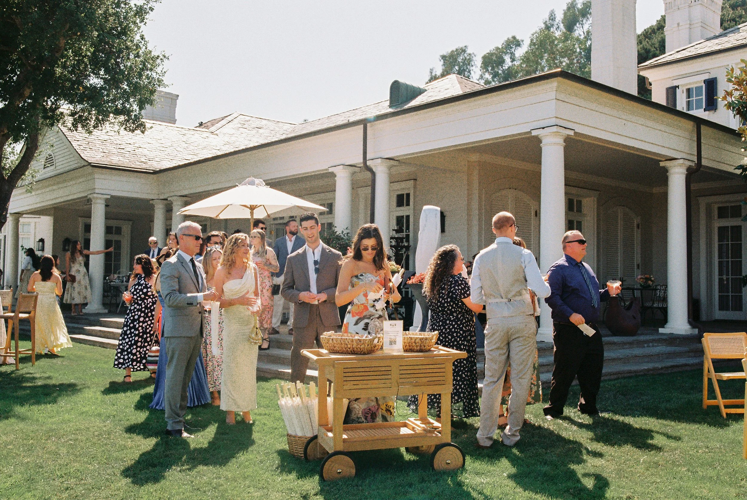 People dressed in formal and casual attire enjoying an outdoor event on a lawn, with a large white house in the background, some holding drinks and engaging in conversations.