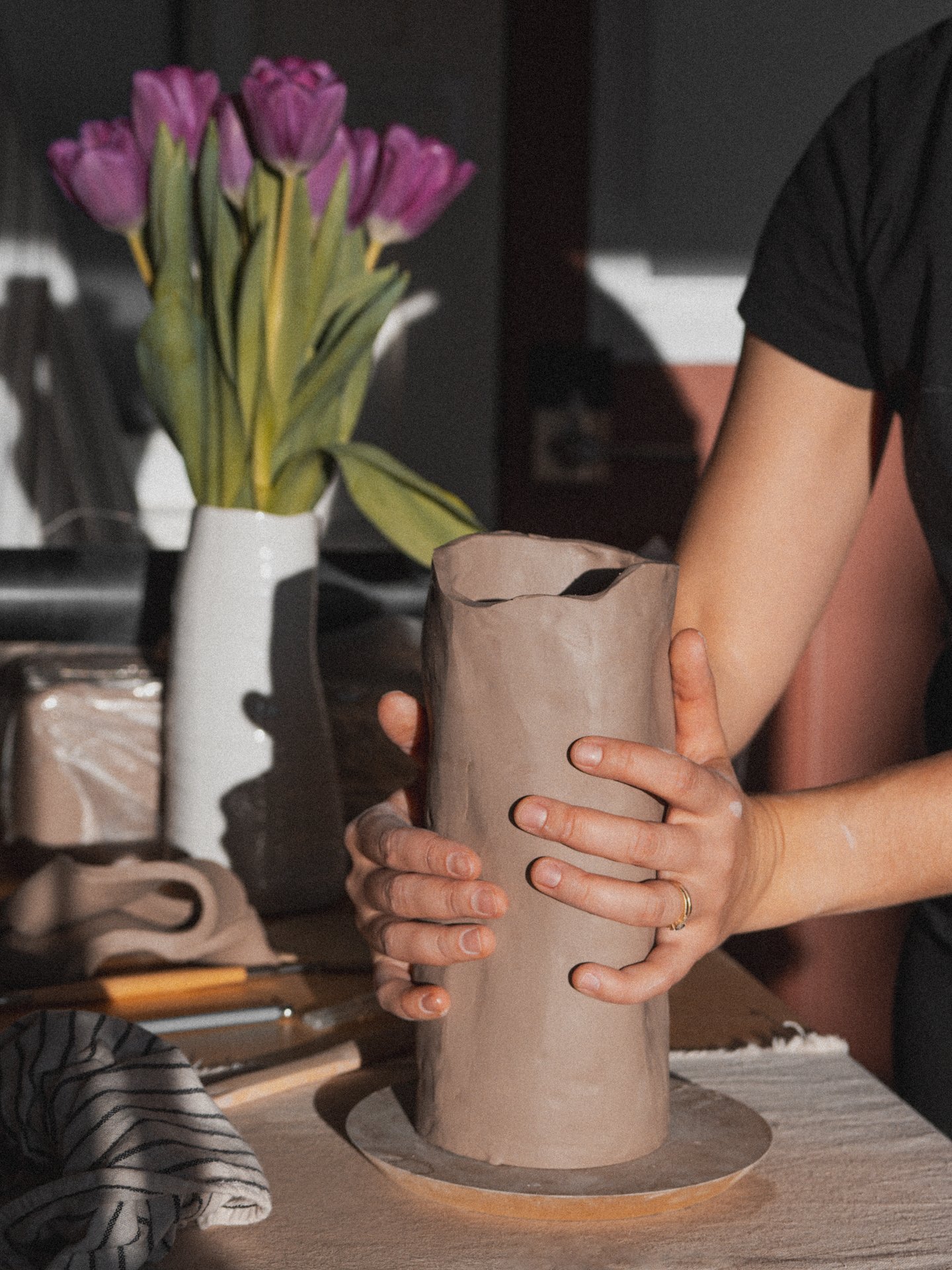 Person shaping a ceramic vase with their hands on a table, with a bouquet of purple tulips in a white vase in the background.