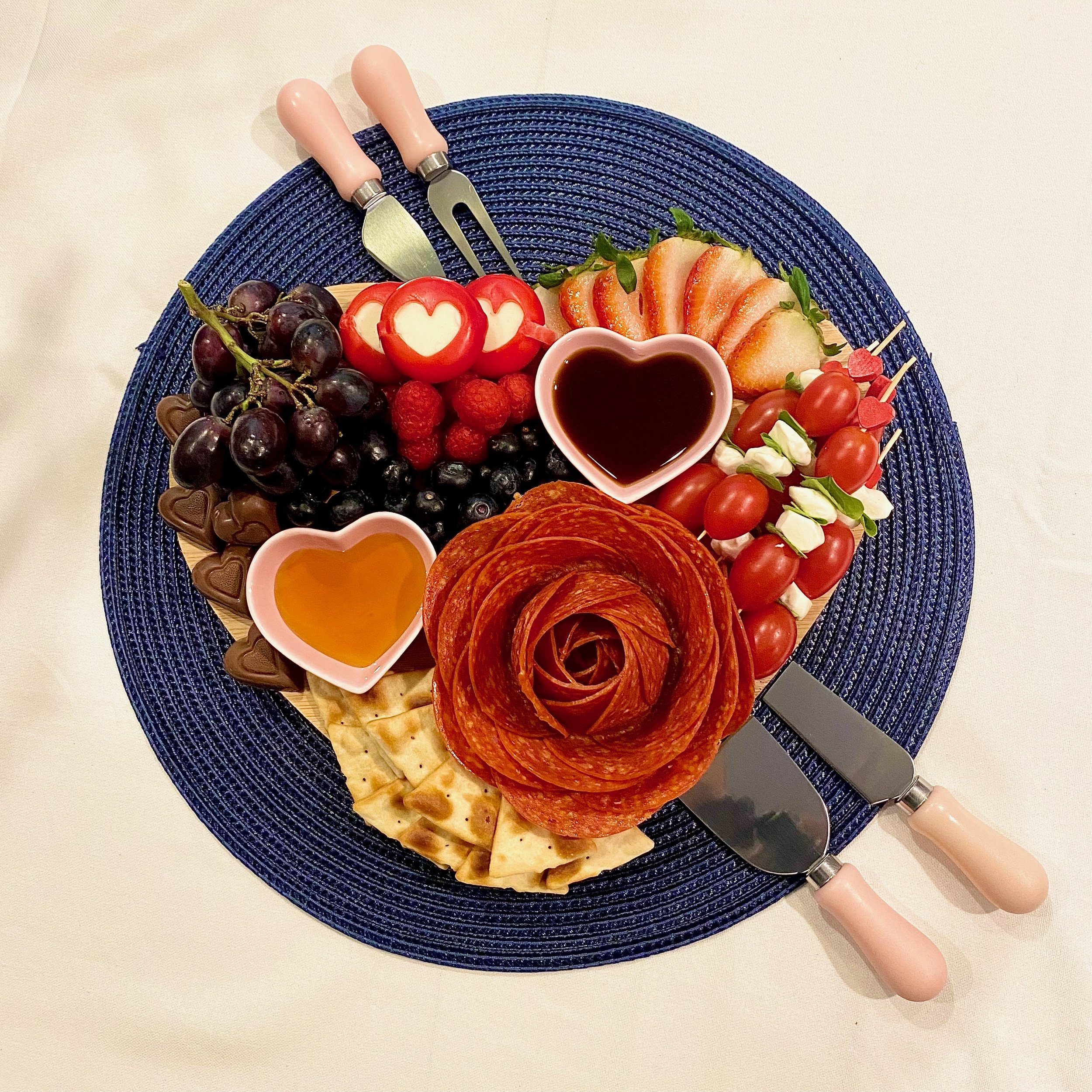 Charcuterie board featuring pepperoni rose, grapes, blueberries, raspberries, sliced strawberries, tomatoes, cheese, crackers, chocolate hearts, and heart-shaped bowls with honey and sauce on a blue mat.