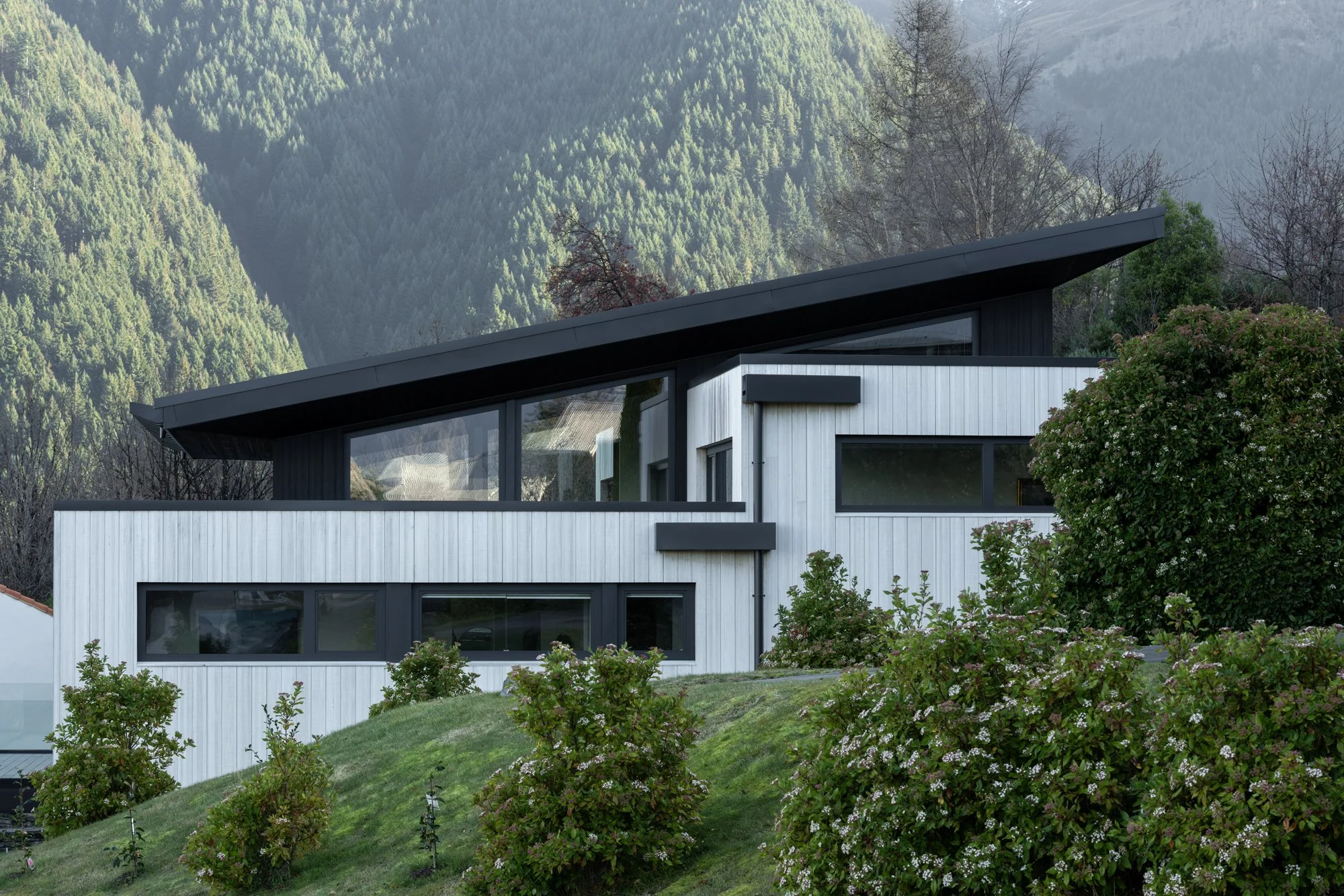 Modern multi-story house with large windows, stone and wood exterior, built into a hillside with mountain in background, clear sky, and landscaped surroundings.
