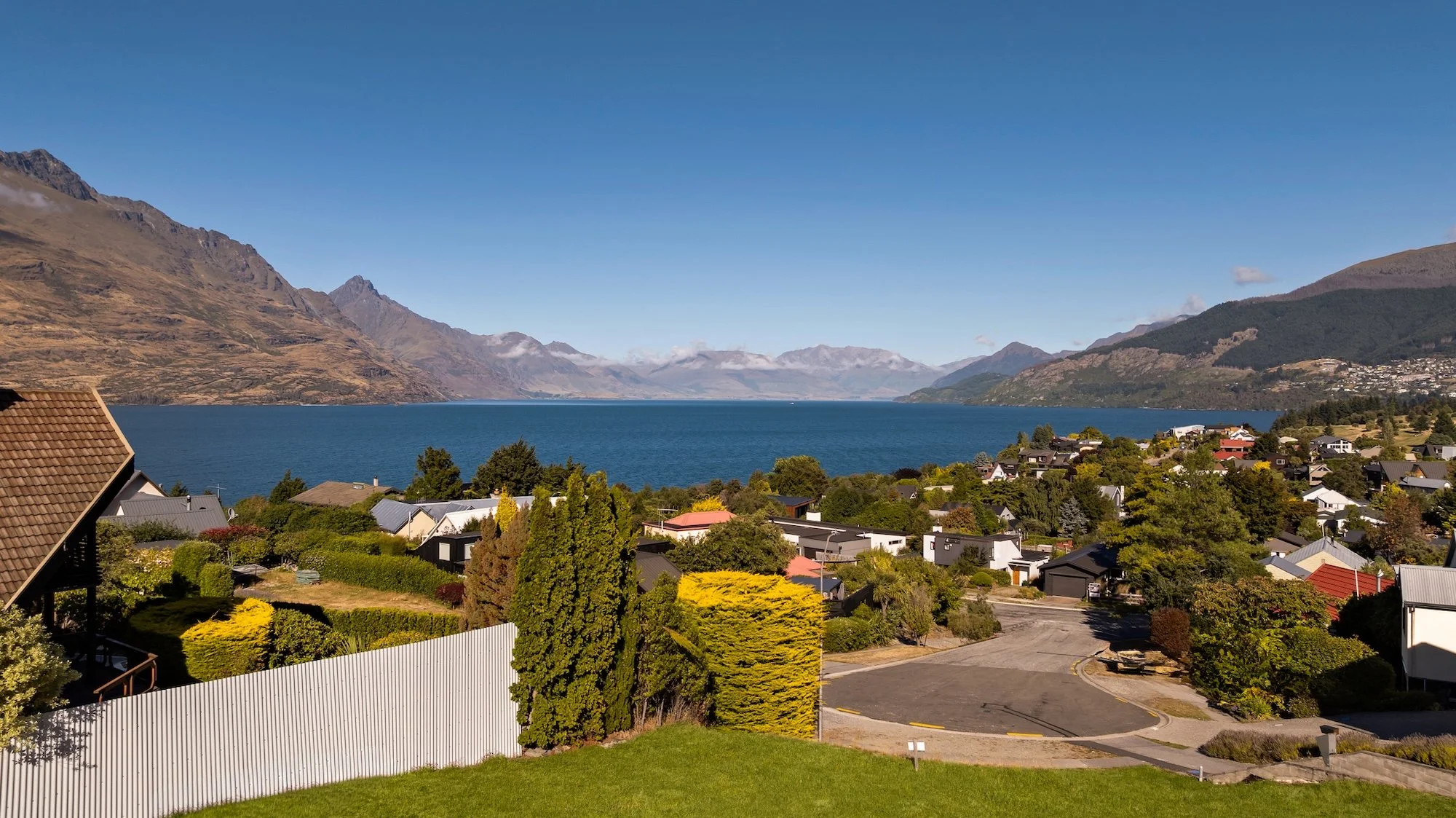 Modern black multi-story house with large windows, situated on a hillside with green grass, bushes, and trees, against a backdrop of a forested mountain and a sunset sky.