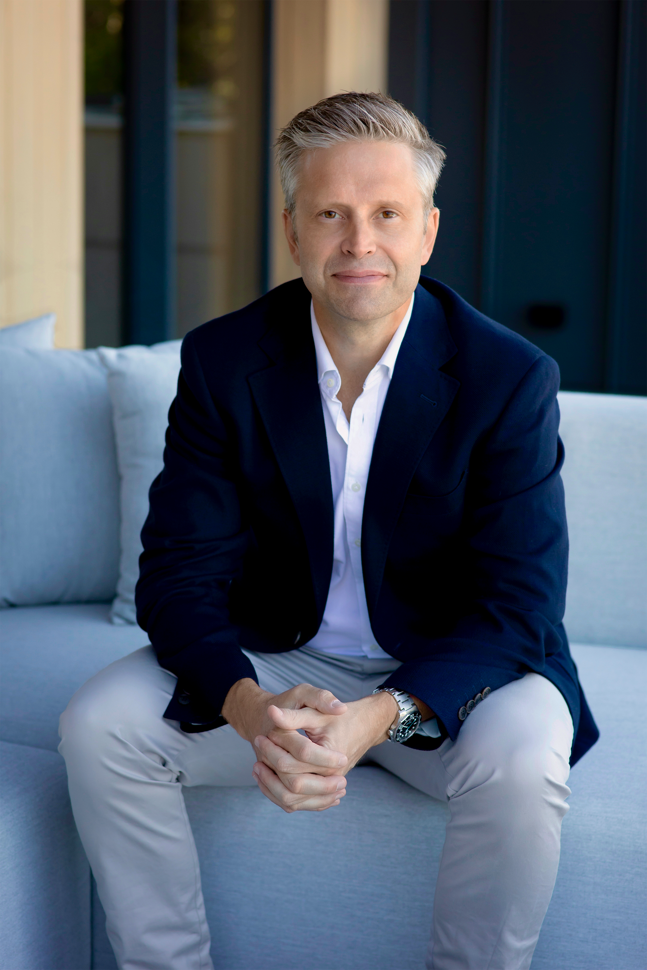 A middle-aged man with silver hair, wearing a dark blue blazer, white shirt, and light-colored pants, sitting casually on a light-colored sofa with his legs apart, hands clasped, and looking at the camera. The background shows a modern indoor setting with wooden and blue elements.