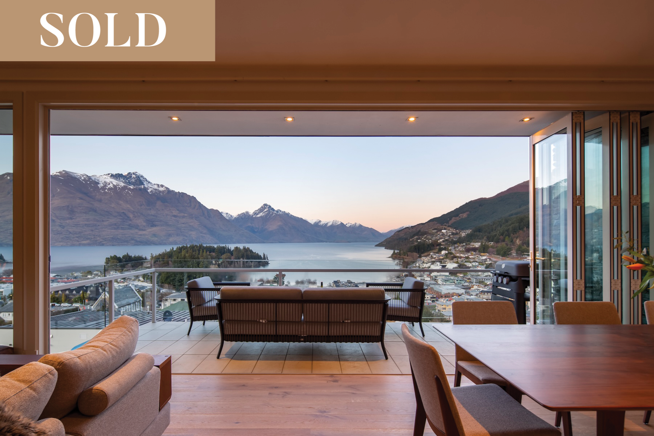 Modern black multi-story house with large windows, situated on a hillside with green grass, bushes, and trees, against a backdrop of a forested mountain and a sunset sky.