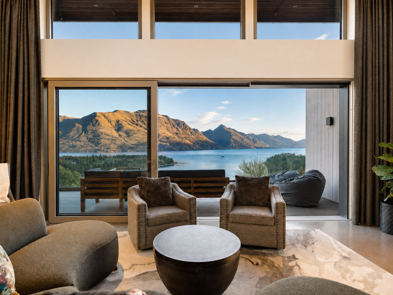 Modern multi-story house with large windows, stone and wood exterior, built into a hillside with mountain in background, clear sky, and landscaped surroundings.
