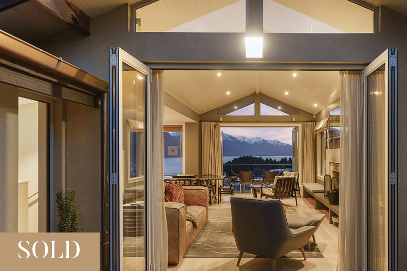 Open glass doors reveal a cozy living room with various chairs, sofas, and a fireplace, featuring large windows with views of snow-capped mountains and a lake at sunset.