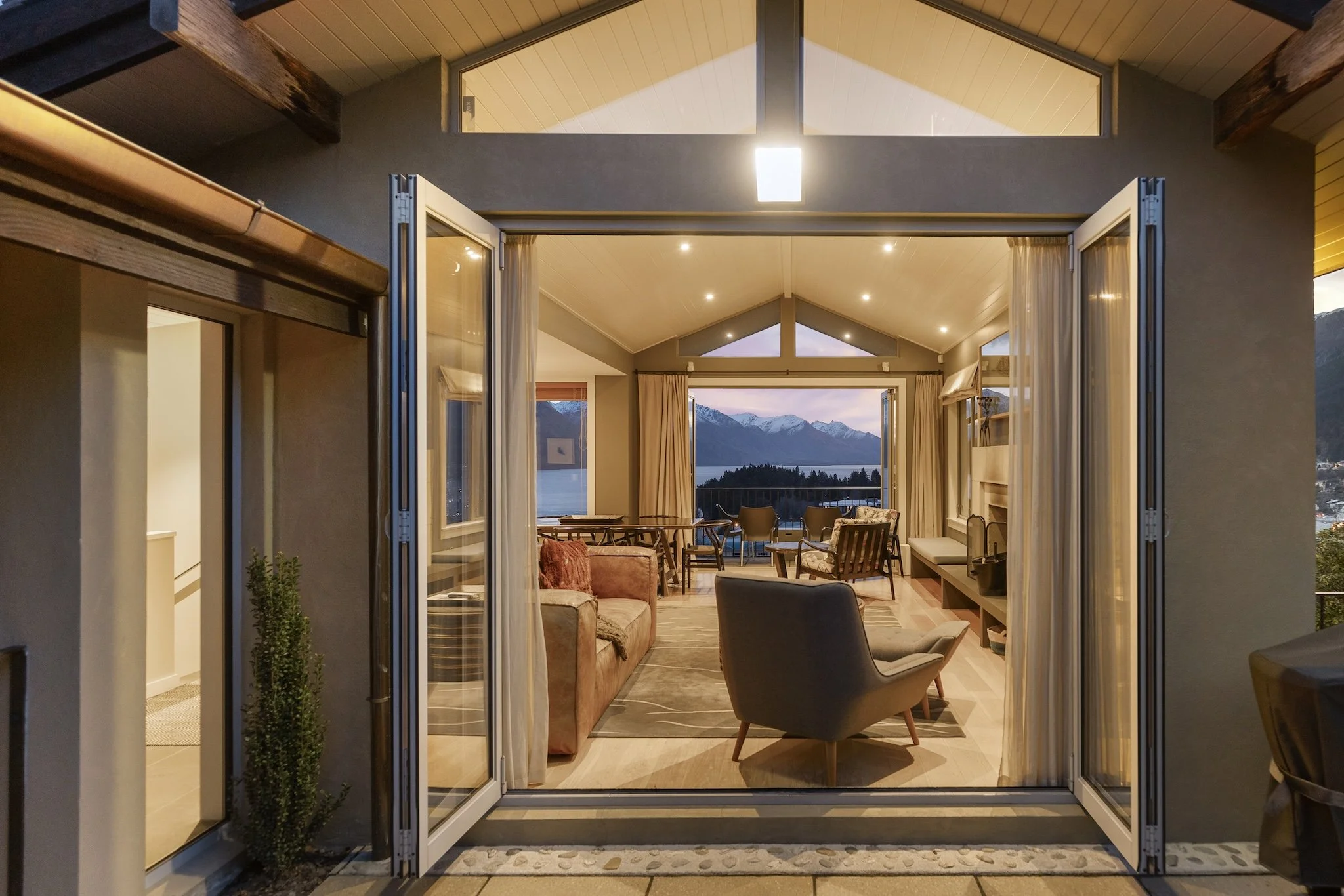 View into a cozy living room with open double doors, overlooking a scenic mountain and lake landscape at sunset.
