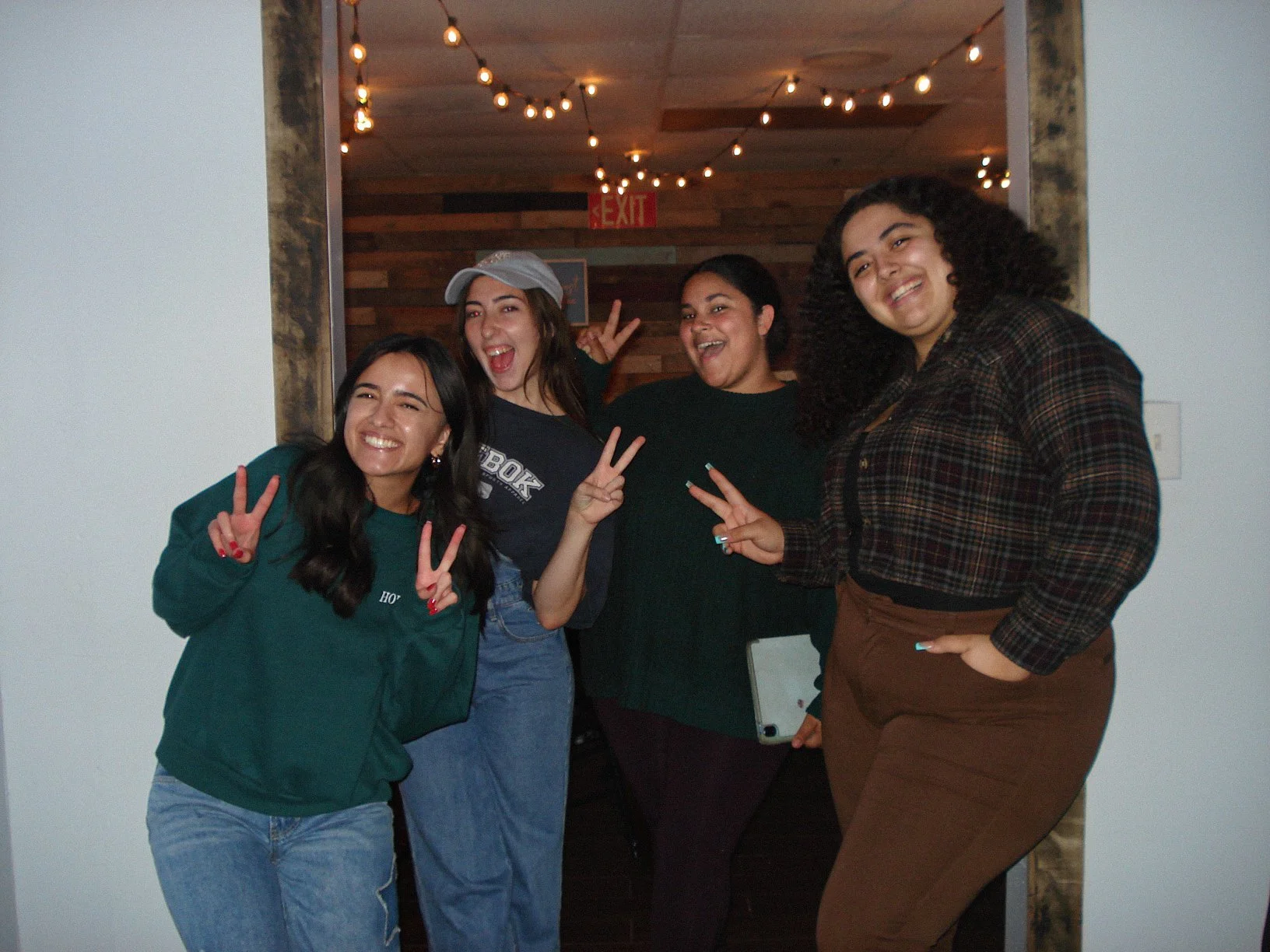 Four women smiling and making peace signs in a doorway, with string lights and wooden decor in the background.
