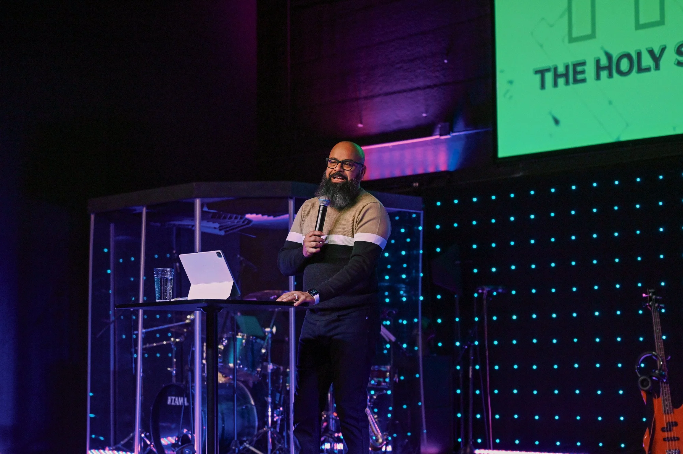 A man with glasses and a beard giving a presentation on stage, holding a microphone, with a laptop and glass of water on a table in front of him. The stage has blue LED lights and a large screen displaying a green slide.