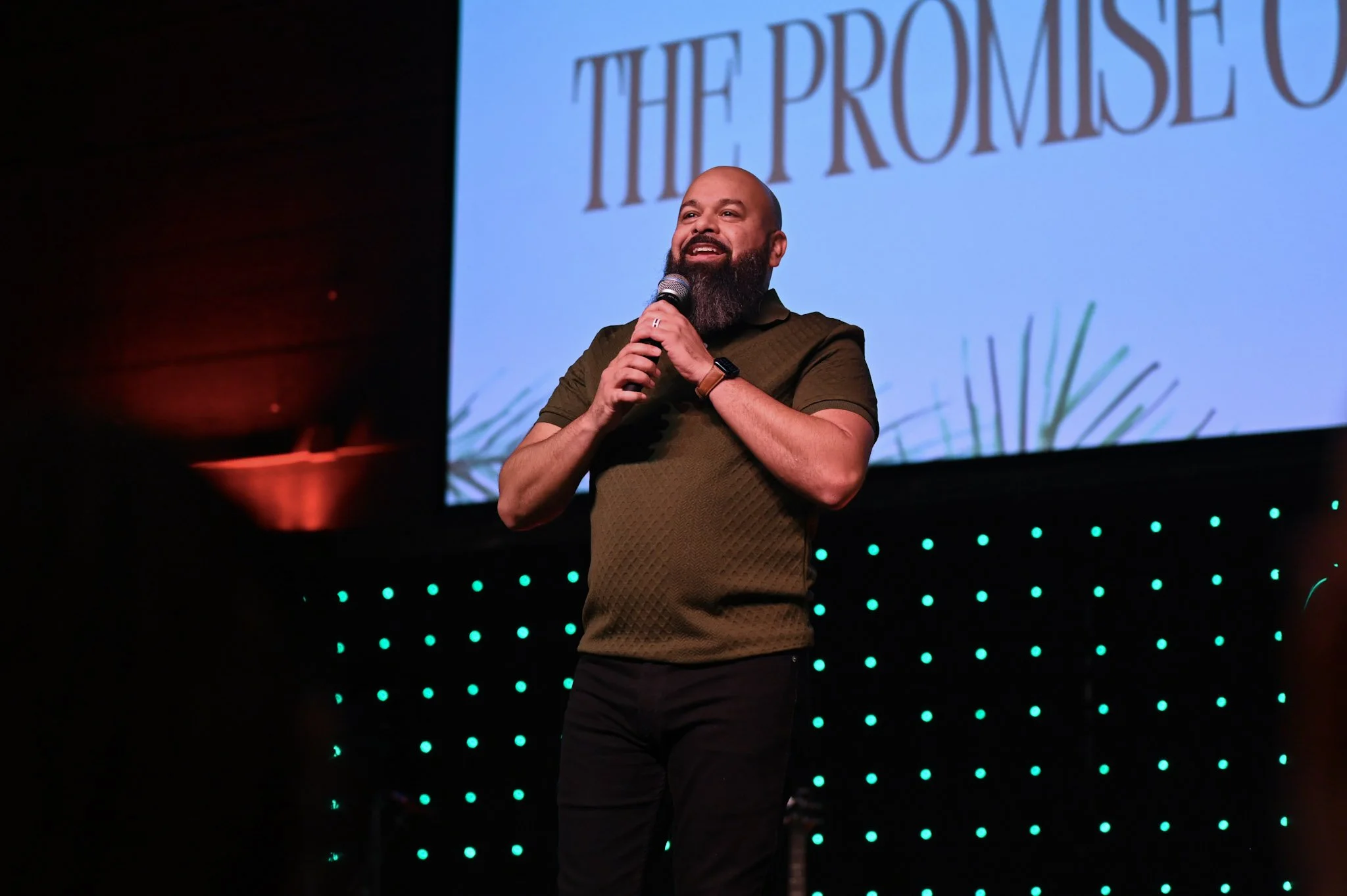 A man with a beard holding a microphone and speaking on stage with a large screen behind him displaying the text 'The Promise of'.