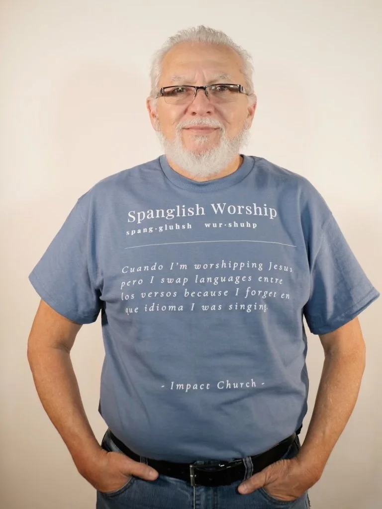 An older man with white hair, a beard, and glasses, wearing a blue T-shirt with text about Spanish worship, standing with hands in his pockets against a plain background.