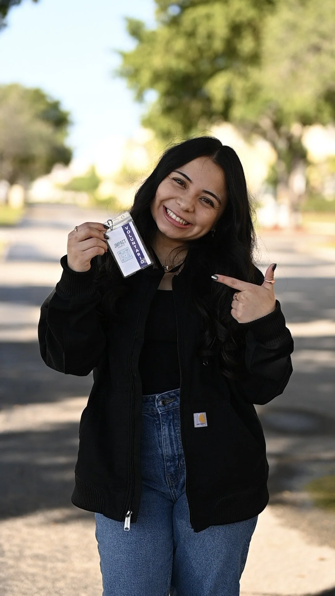 A woman smiling outdoors, holding a security badge in her left hand and pointing at it with her right finger. She has long dark hair, wearing a black jacket and jeans, with a background of trees and a clear sky.