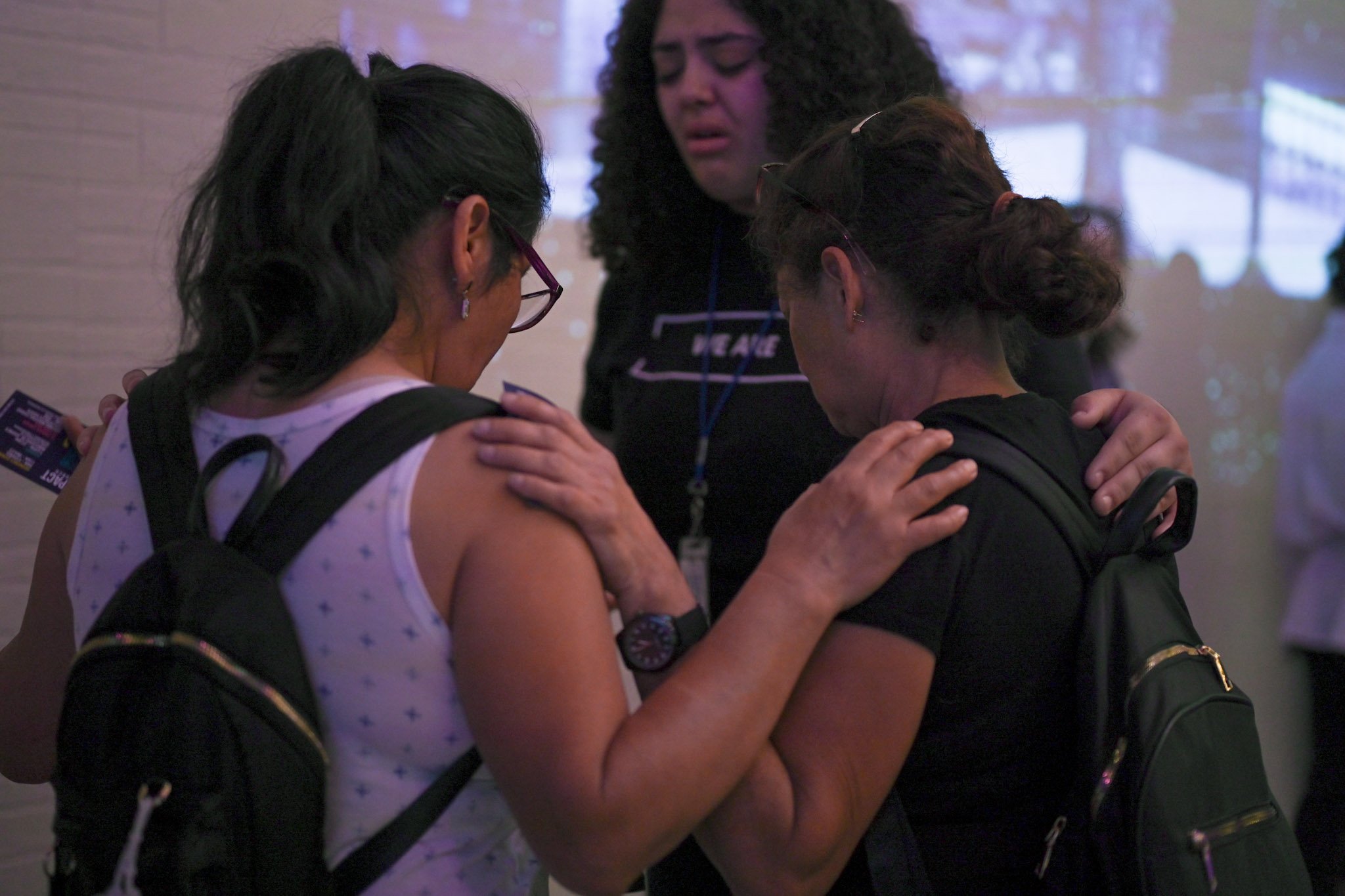 Three women pray together, with two women wearing backpacks and bowing their heads, one woman has her hand on the other's shoulder, in a moment of prayer or reflection.