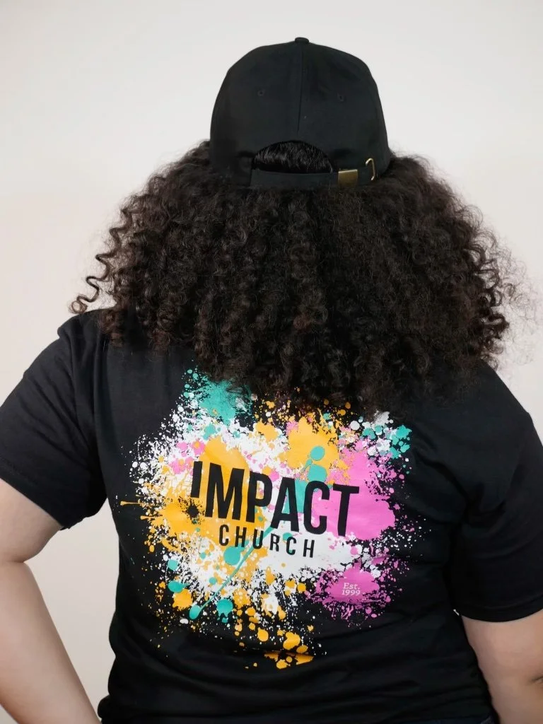 Person wearing a black cap and a black T-shirt with colorful splash paint design and the words 'Impact Church' on the back.