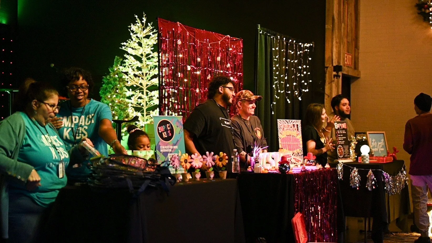 Group of people standing behind decorated tables with Christmas lights and trees in the background at an indoor festive event.