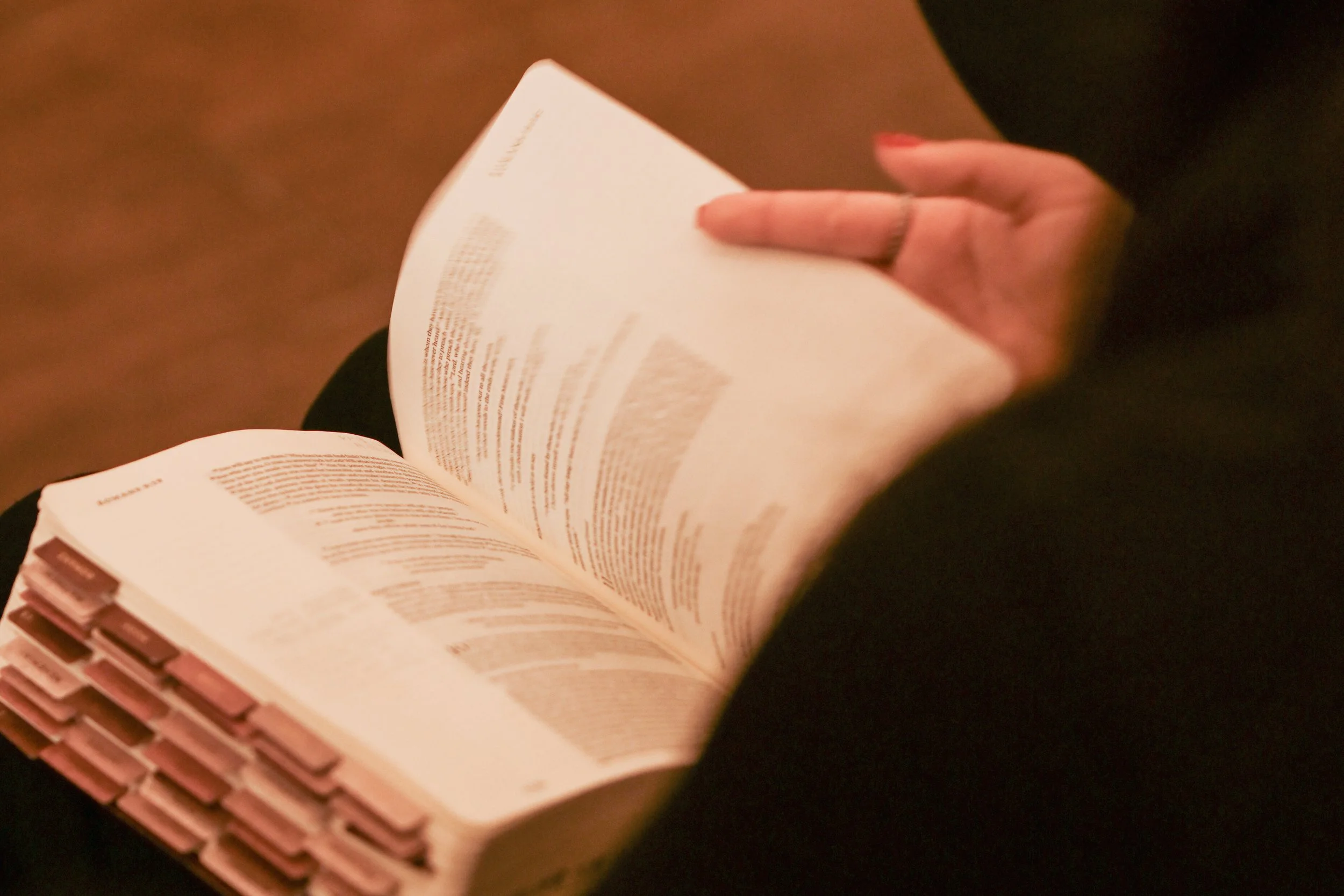 Person holding an open book with text, sitting on a chair or bench, with a brown floor and wearing a black sleeve.