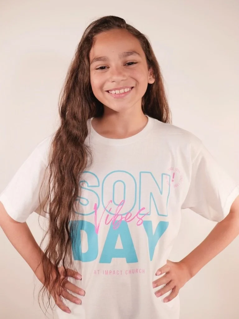 A young girl with long, wavy brown hair smiling and posing with her hands on her hips, wearing a white T-shirt that says "SON Day Vibes" and references Impact Church.