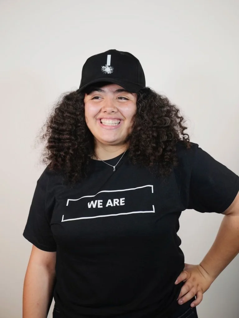A woman with curly hair smiling, wearing a black cap and a black t-shirt with 'WE ARE' text, standing against a plain background.