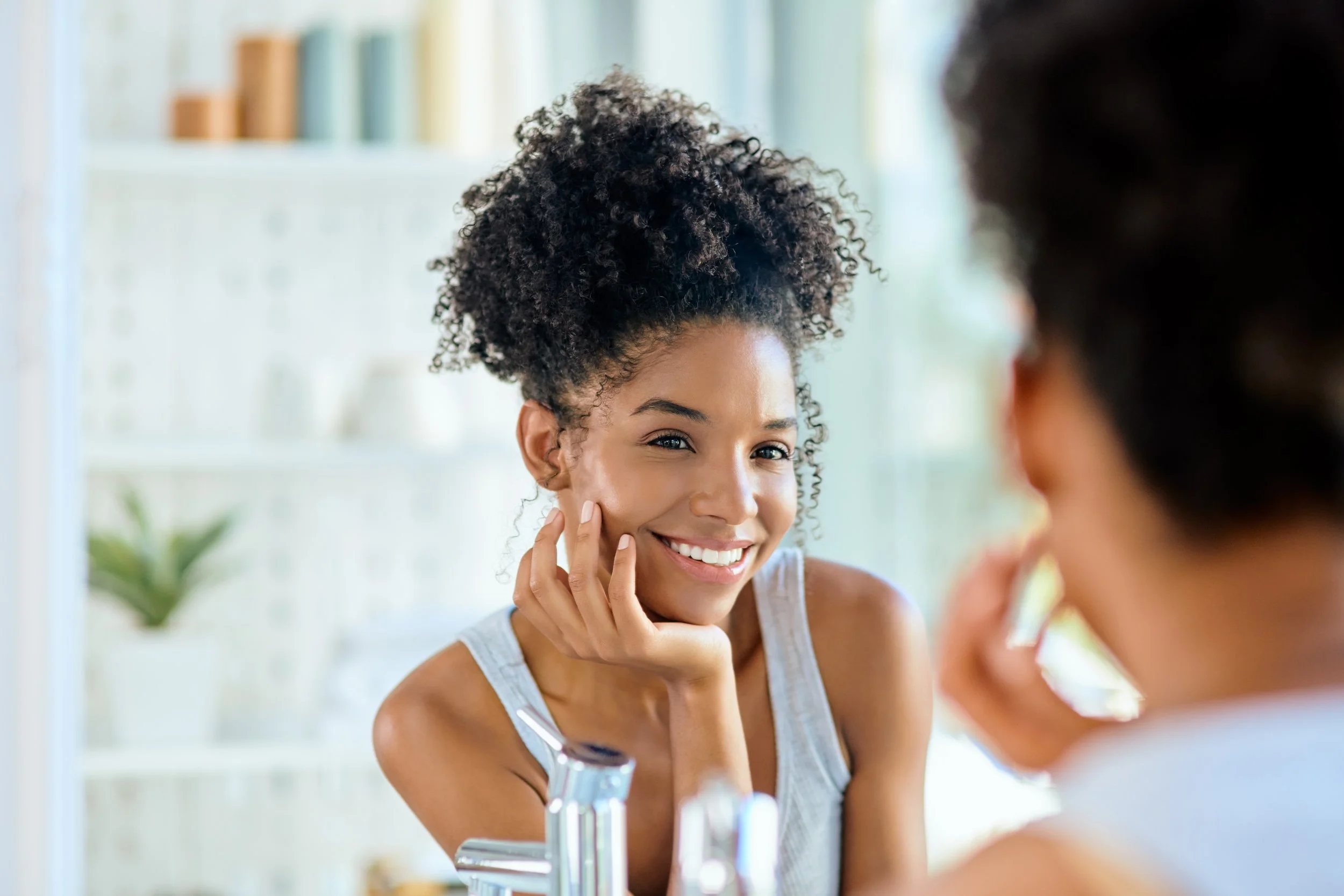 Woman with curly hair smiling while looking at her skin in a mirror during a skincare consultation.