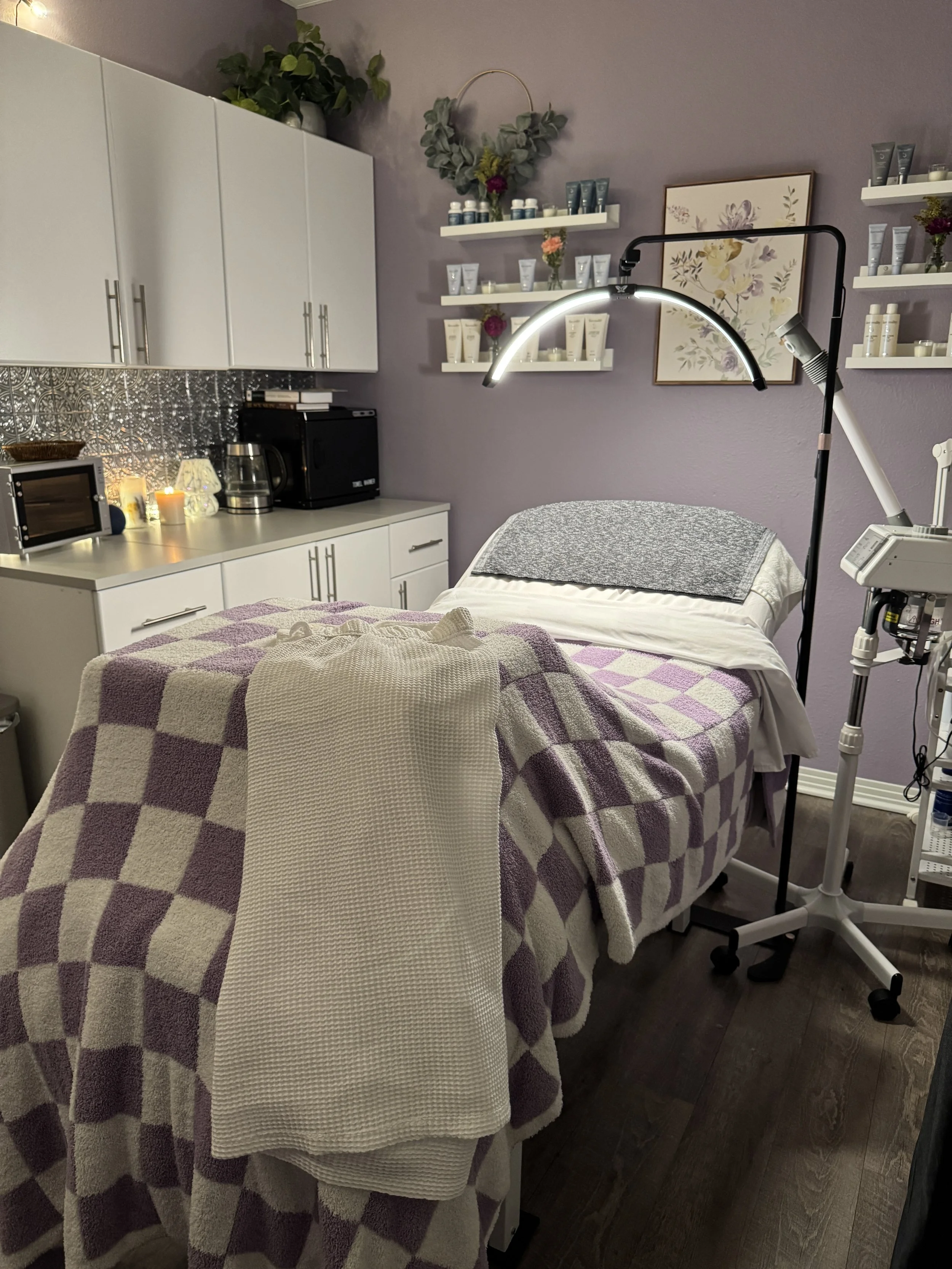Skincare studio treatment room at Cherish Beauty Aesthetics with a facial treatment bed, towel warmer, skincare equipment, candles, and shelves of professional skincare products.