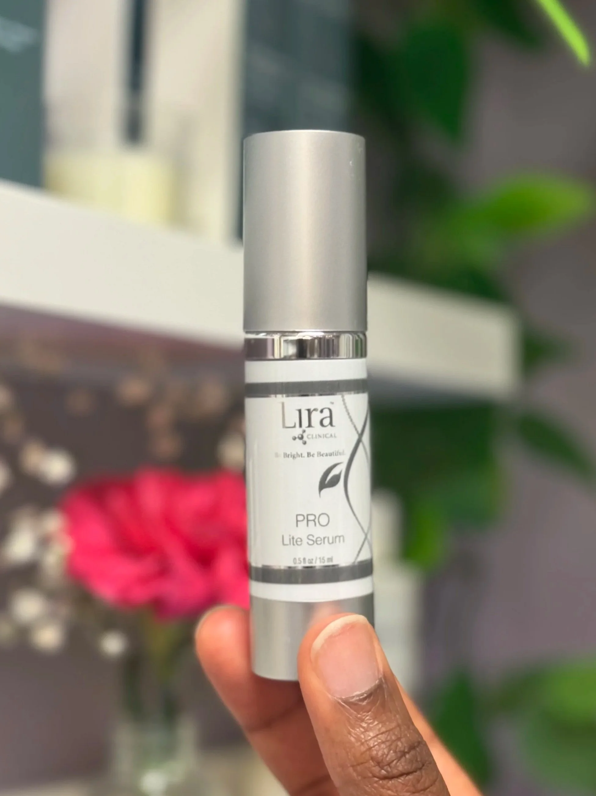 A hand holding a white bottle labeled 'Lira Clinical PRO Lite Serum' with a silver cap, with blurred pink flowers and green foliage in the background.