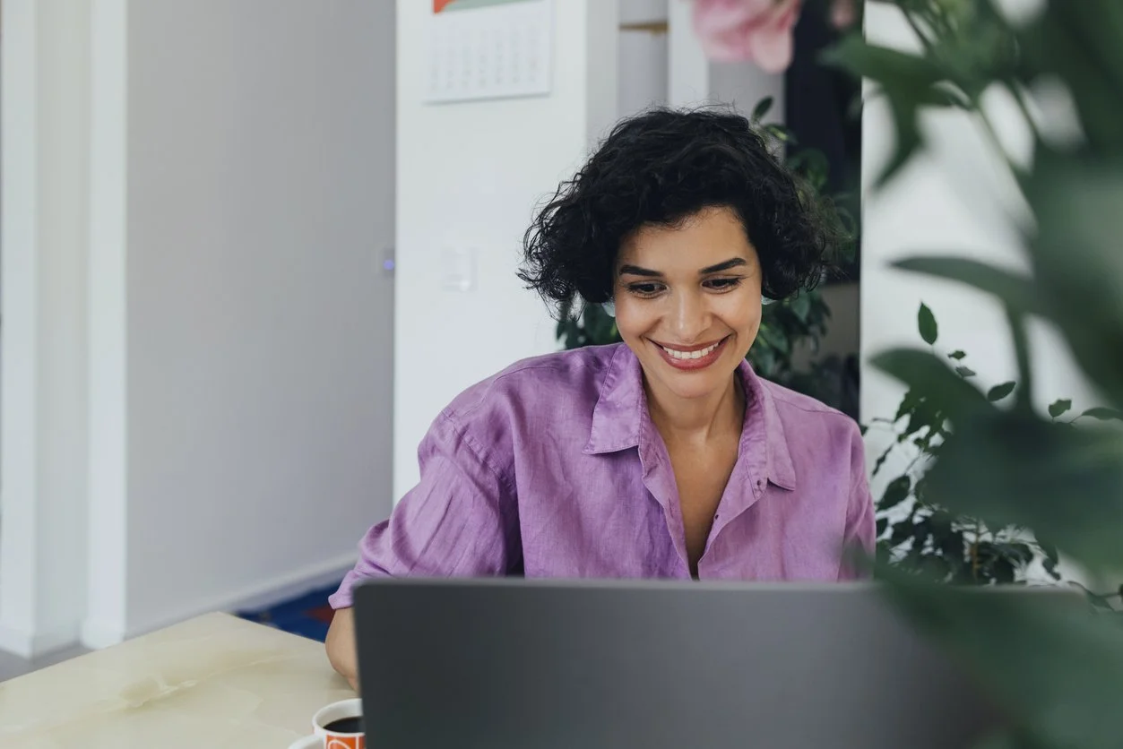 Woman with curly black hair and a purple shirt working on a laptop at a desk, smiling. There are plants and a cup of coffee nearby, in a bright office space.