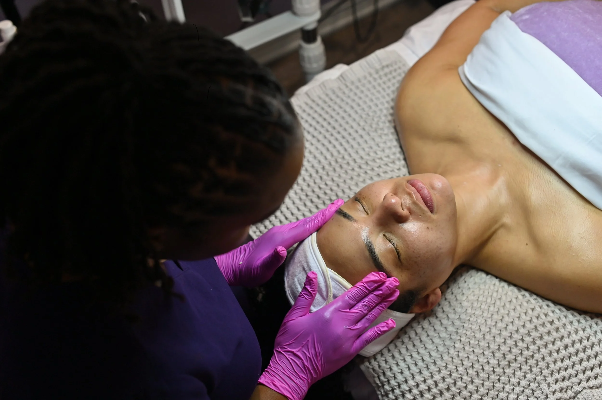 A woman receiving a facial treatment from a skincare professional wearing pink gloves in a spa or skincare clinic.