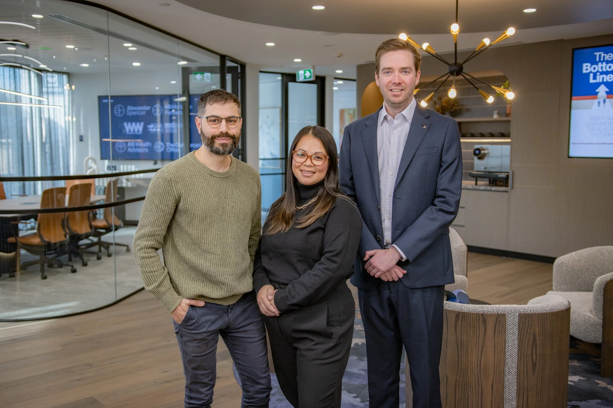 Three professionals, two men and one woman, standing together in a modern office lounge area, smiling at the camera.